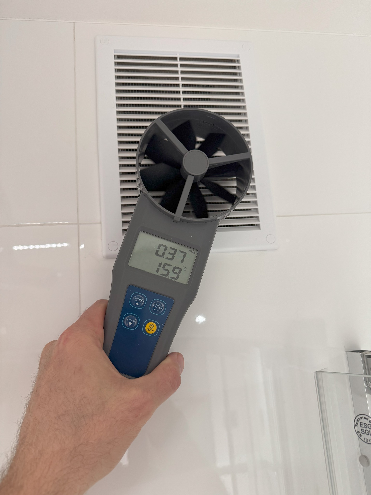 Pomiar prędkości przepływu powietrza anemometrem w kratce wentylacyjnej, wskazania na wyświetlaczu 0.37 m/s i 15.9 stopni Celsjusza.