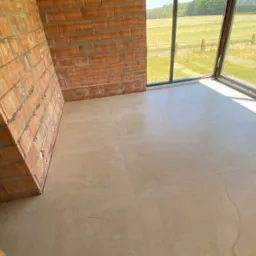 Jasne płytki podłogowe w surowym wnętrzu z ceglaną ścianą i dużym oknem z widokiem na zieleń. Widoczne białe kable. Neutralna kolorystyka.