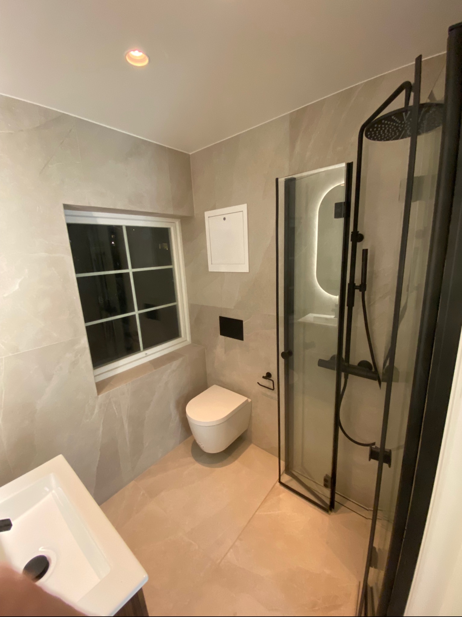 Nowoczesna łazienka z beżowymi płytkami, kabiną prysznicową z czarnymi ramami, wiszącą toaletą i oknem z białymi szprosami.