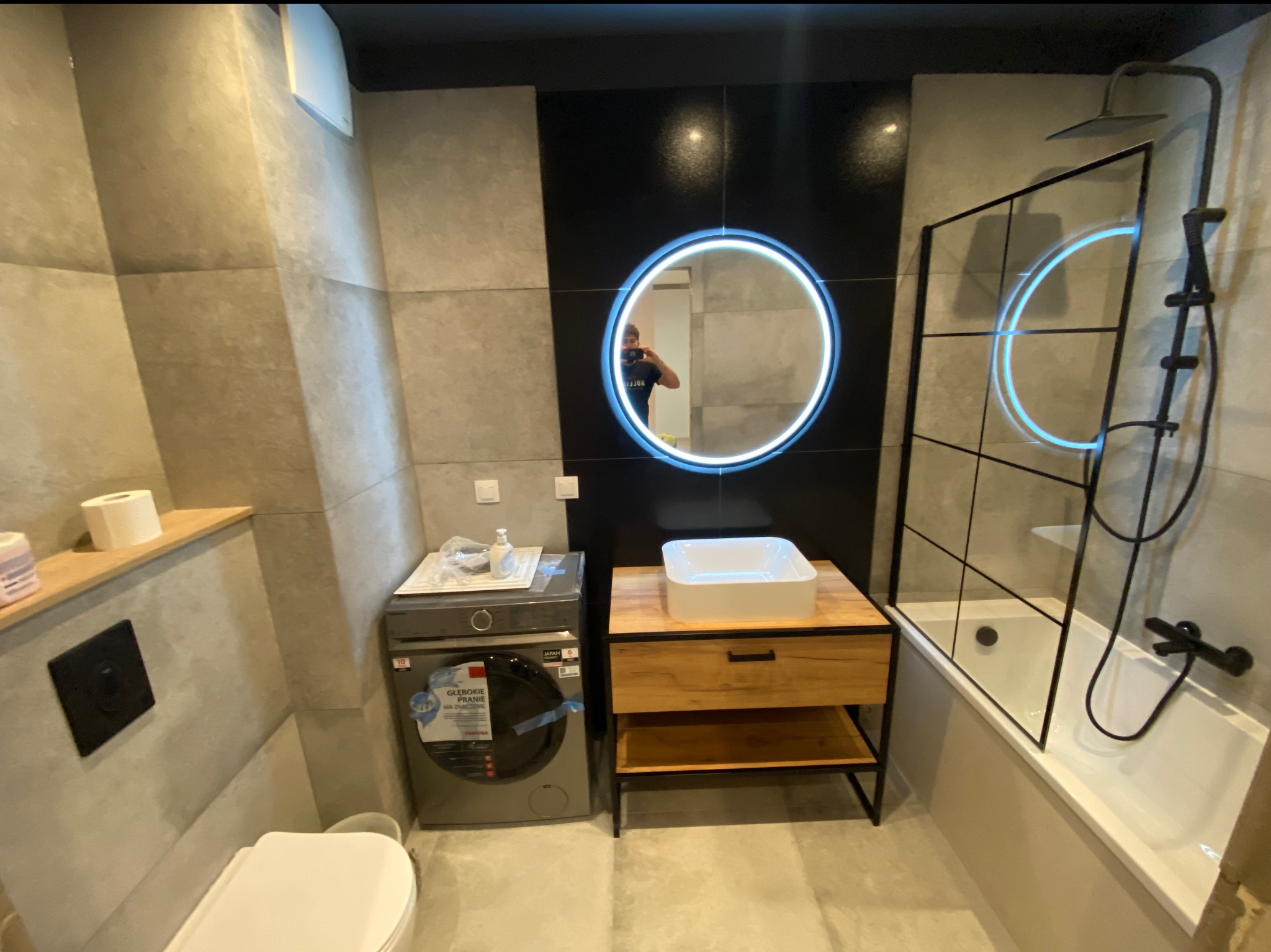 Nowoczesna łazienka z szarymi płytkami, okrągłym lustrem z podświetleniem LED, pralką i wanną z czarną armaturą oraz szklanym parawanem.