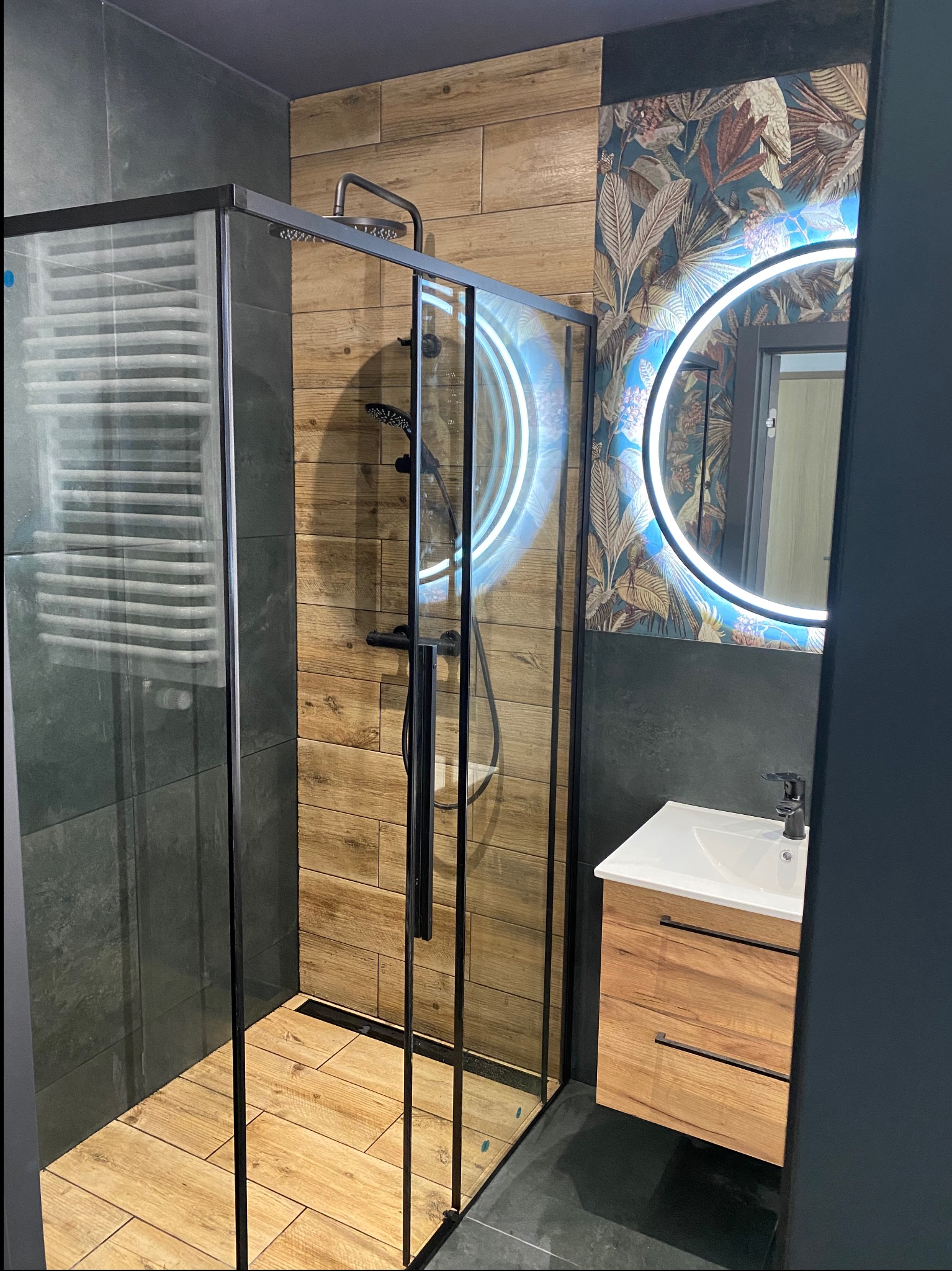 Nowoczesna łazienka z kabiną prysznicową typu walk-in z czarnymi profilami, drewnopodobnymi płytkami na ścianach i podłodze, okrągłym lustrem z podświetleniem LED oraz szafką pod umywalką...