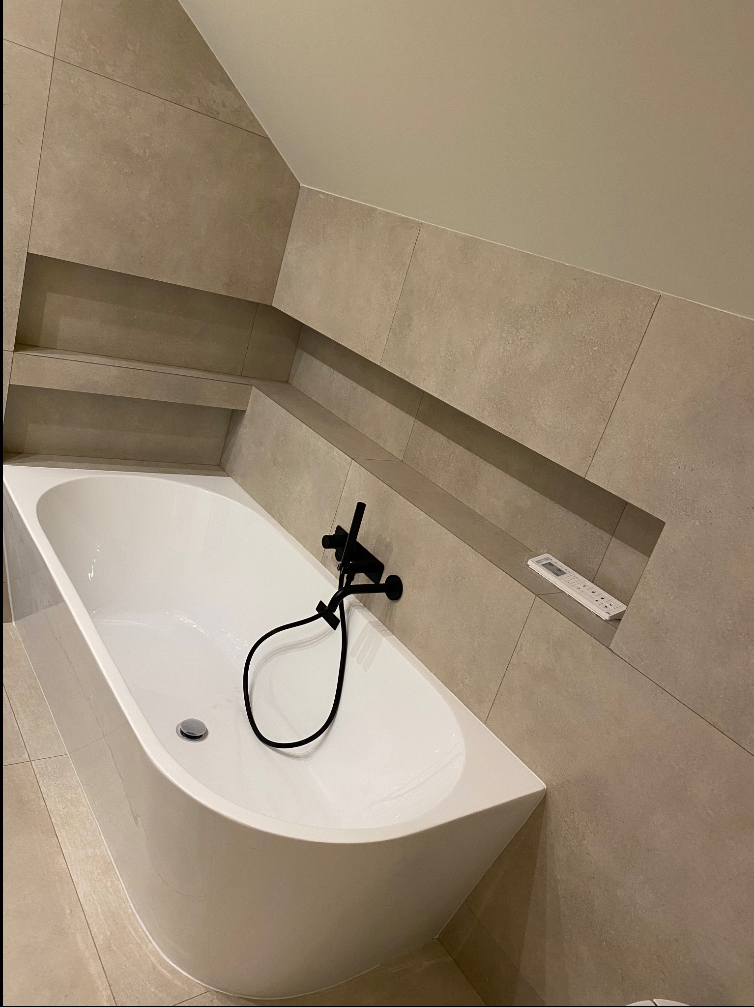 Nowoczesna łazienka w odcieniach beżu z wolnostojącą wanną, czarną armaturą i wnęką w ścianie, w której leży pilot.