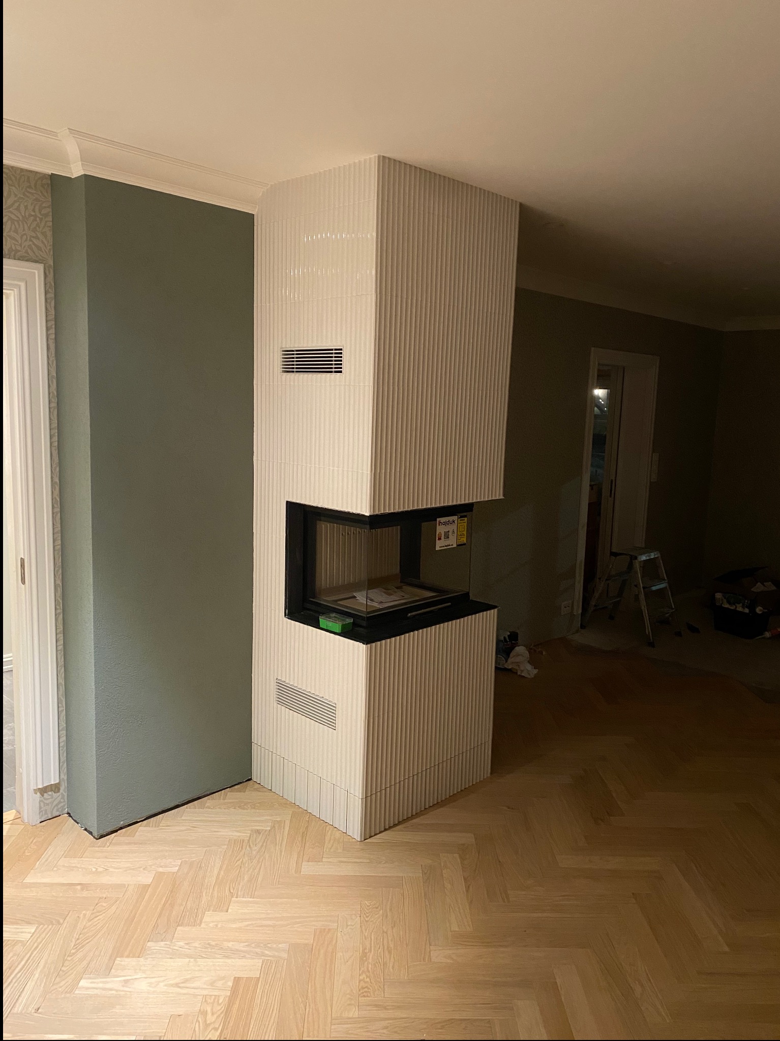 Nowoczesny kominek obłożony białymi, ryflowanymi płytkami ceramicznymi, z czarnym wkładem kominkowym, w trakcie wykończenia, widoczny fragment parkietu i drabina w tle.