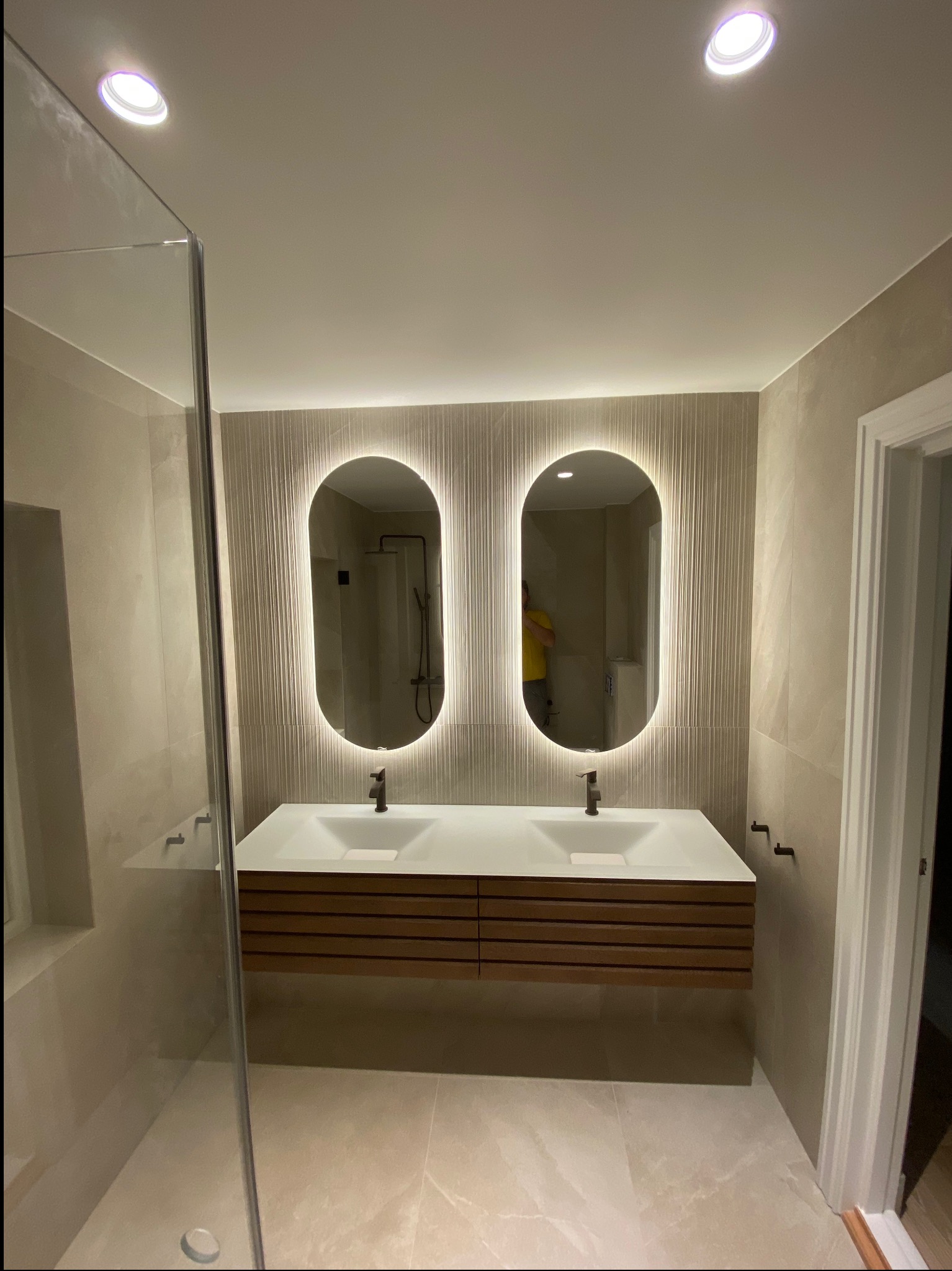Nowoczesna łazienka z dwoma owalnymi lustrami z podświetleniem LED, wisząca szafka z umywalką z drewnianymi lamelami, kabina prysznicowa ze szklaną ścianką i beżowe płytki na ścianach i podłodze.
