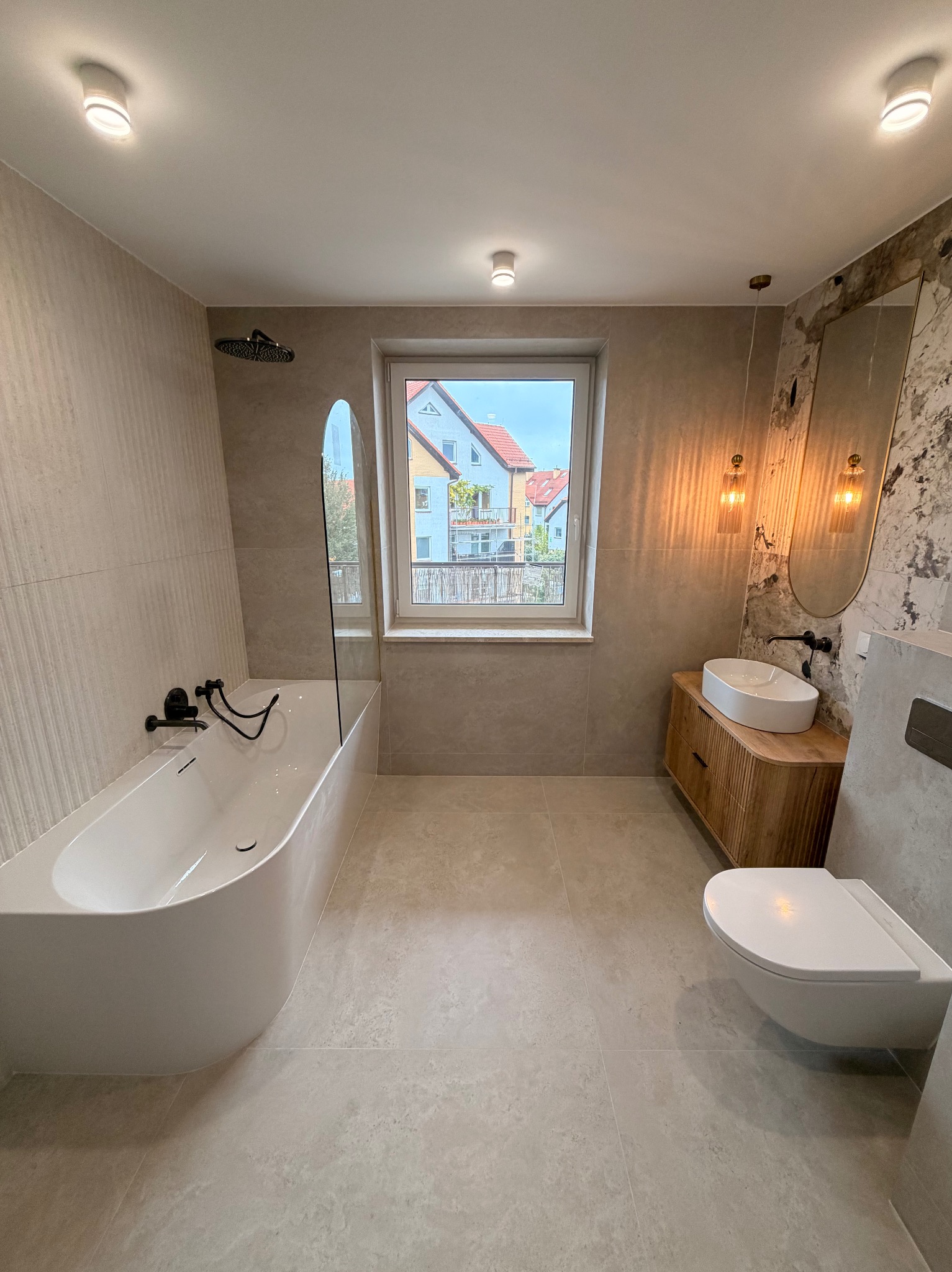 Nowoczesna łazienka z wanną, umywalką na drewnianej szafce i toaletą podwieszaną. Jasne płytki i okno z widokiem na budynki.