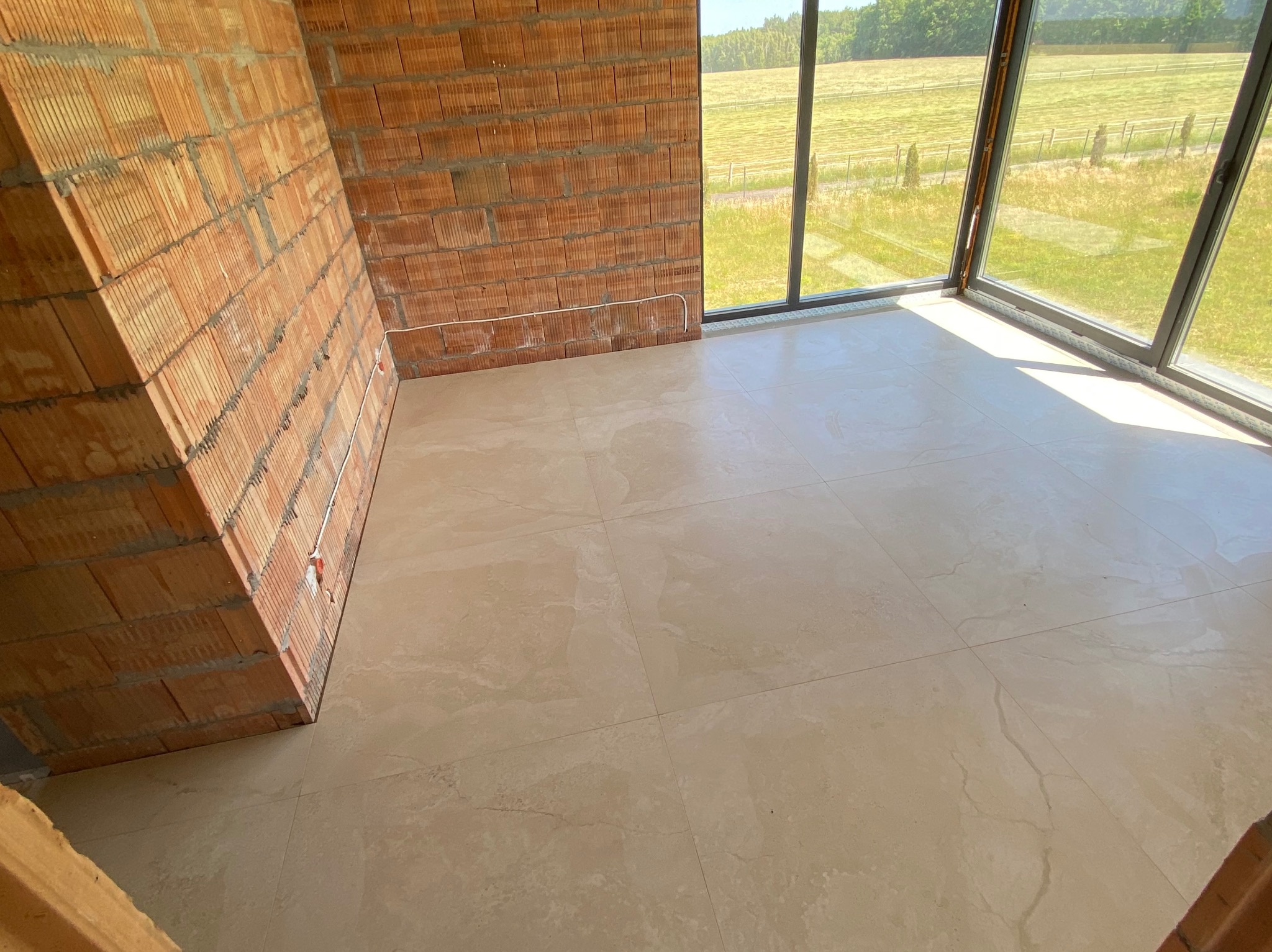 Jasne płytki podłogowe w surowym wnętrzu z ceglaną ścianą i dużym oknem z widokiem na zieleń. Widoczne białe kable. Neutralna kolorystyka.