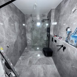 Rafal Chabiniak - Szara łazienka po remoncie z kabiną prysznicową typu walk-in, czarną armaturą i wiszącą toaletą. Na ścianach i podłodze płytki imitujące marmur. Widoczne narzędzia.