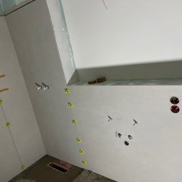Rafal Chabiniak - Ściany w trakcie montażu płyt gipsowo-kartonowych w łazience, widoczne profile, żółte krzyżyki poziomujące i instalacje elektryczne. Surowy stan wykończenia.