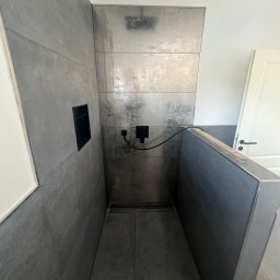 Rafal Chabiniak - Nowoczesna kabina prysznicowa z szarymi płytkami imitującymi beton, czarną armaturą i wnęką. Minimalistyczny design, surowy styl, widoczny odpływ liniowy.