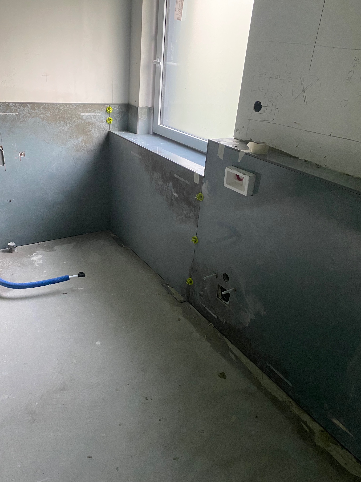 Łazienka w trakcie remontu: szare płytki częściowo ułożone, widoczne krzyżyki glazurnicze, przygotowane punkty instalacyjne i odręczne notatki na ścianie.