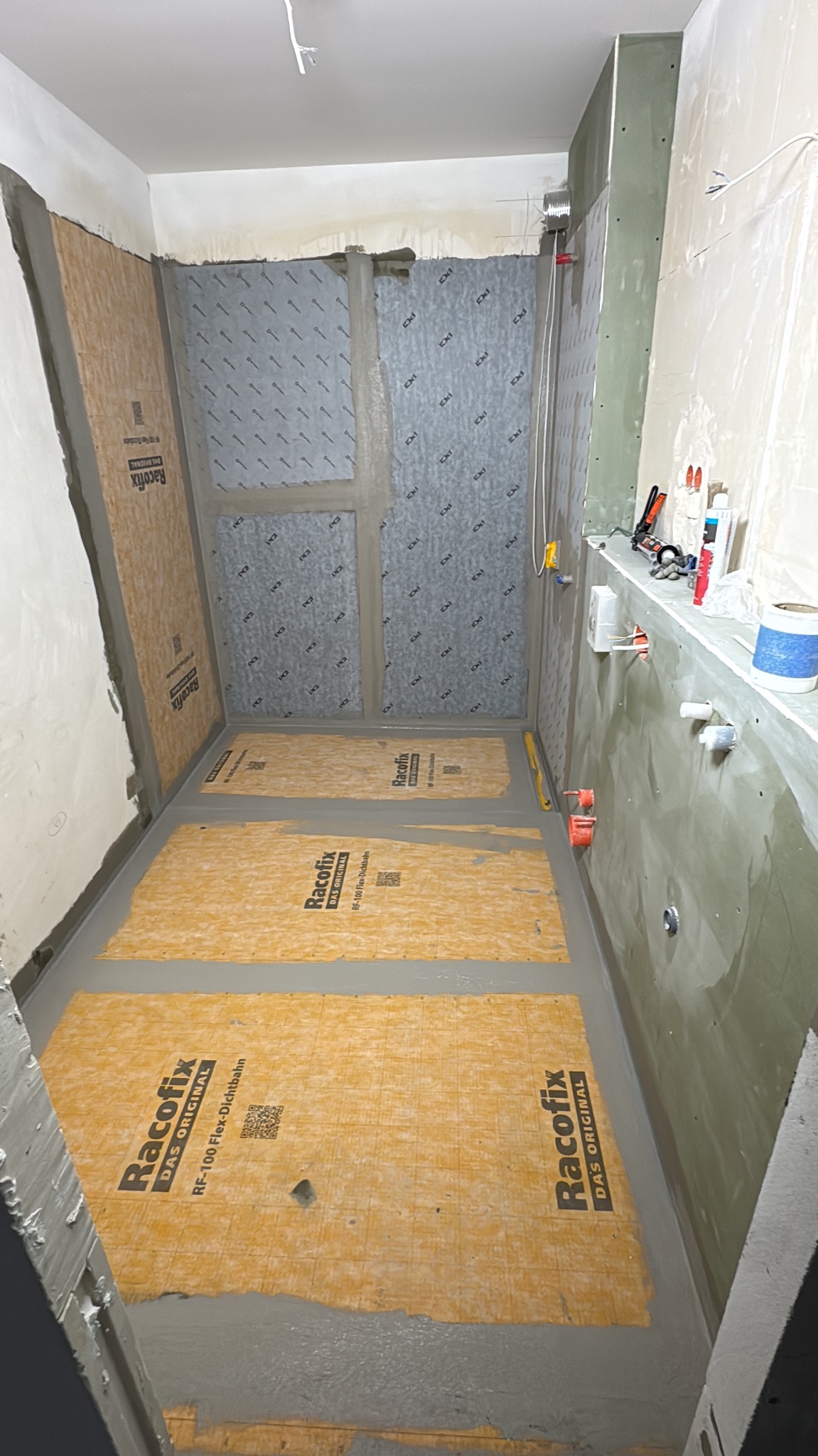 Przygotowanie do hydroizolacji w łazience: folia w płynie na podłodze i ścianach, widoczne narzędzia i instalacje. Etap prac przed położeniem płytek.