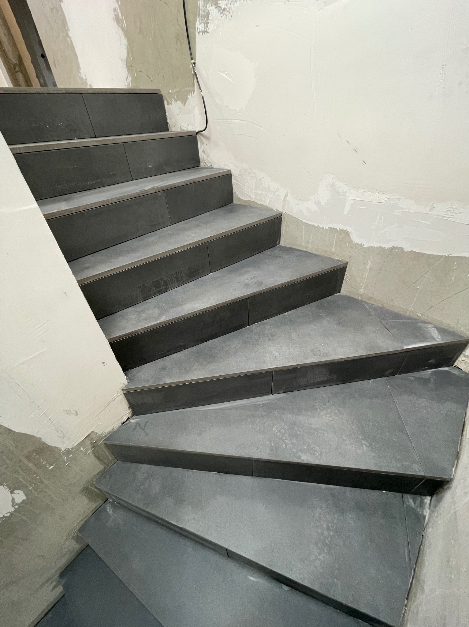 Wykończone schody z szarych płytek, widoczne ślady prac wykończeniowych na ścianach. Ujęcie z dołu, podkreślające geometrię stopni.
