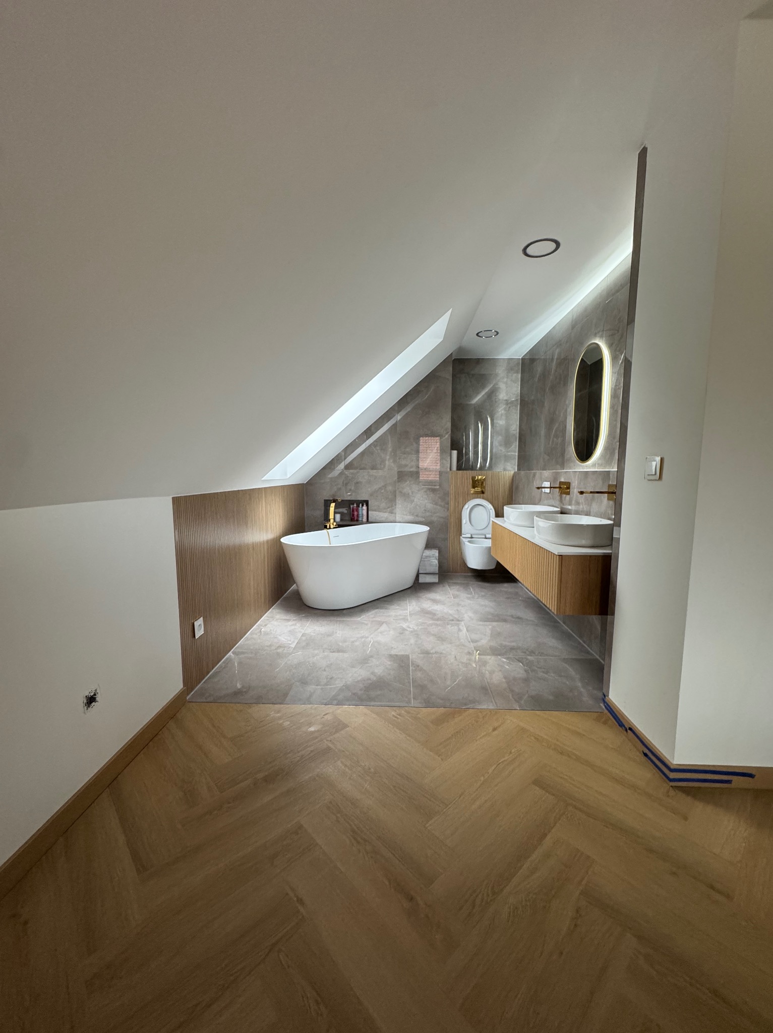 Nowoczesna łazienka na poddaszu z wolnostojącą wanną, marmurowymi płytkami i drewnianymi akcentami. Umywalki nablatowe i podświetlane lustro dodają elegancji.