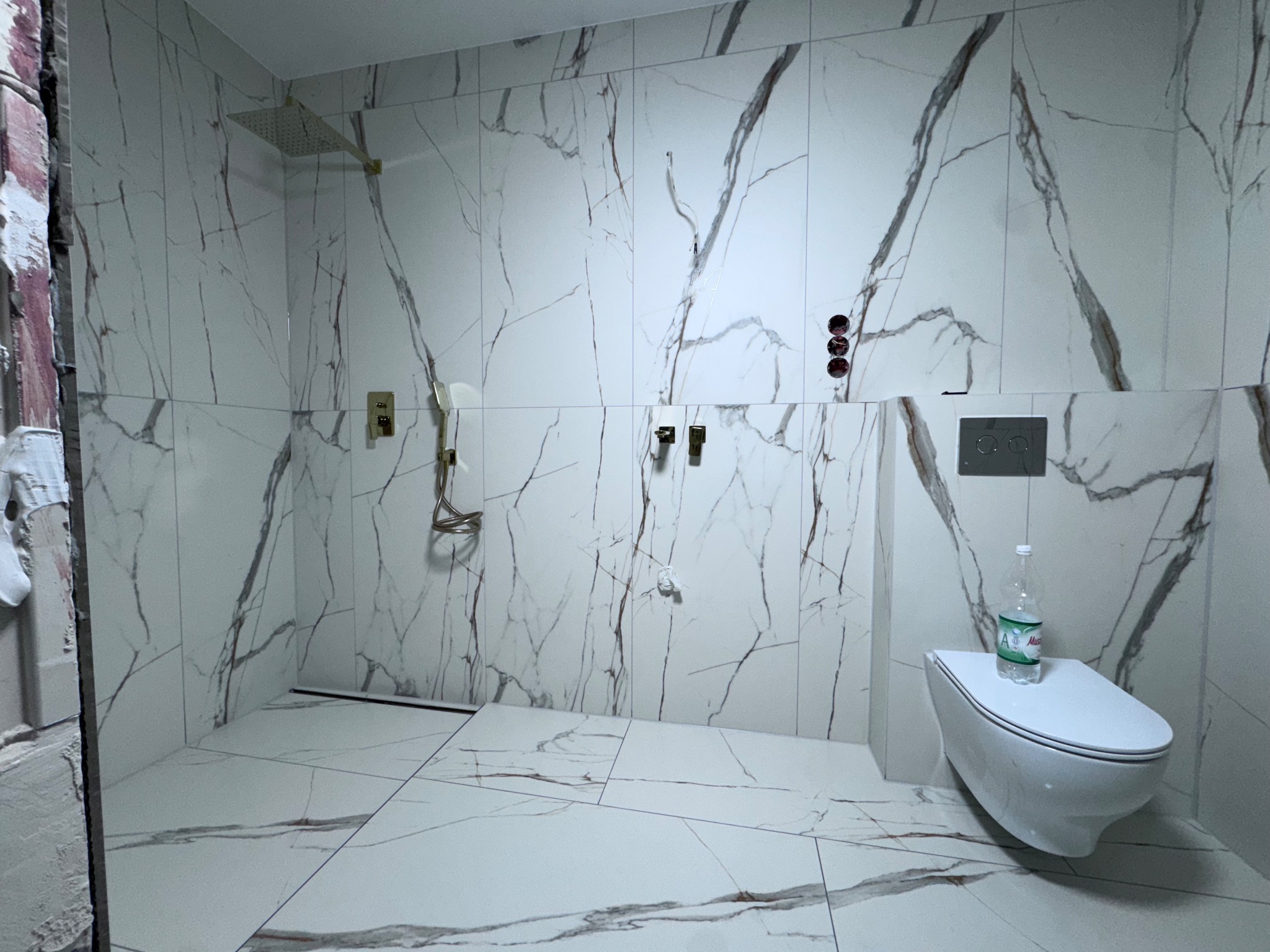 Łazienka w trakcie wykończenia: białe płytki imitujące marmur na ścianach i podłodze, wisząca toaleta z butelką wody, widoczne elementy instalacji prysznicowej.
