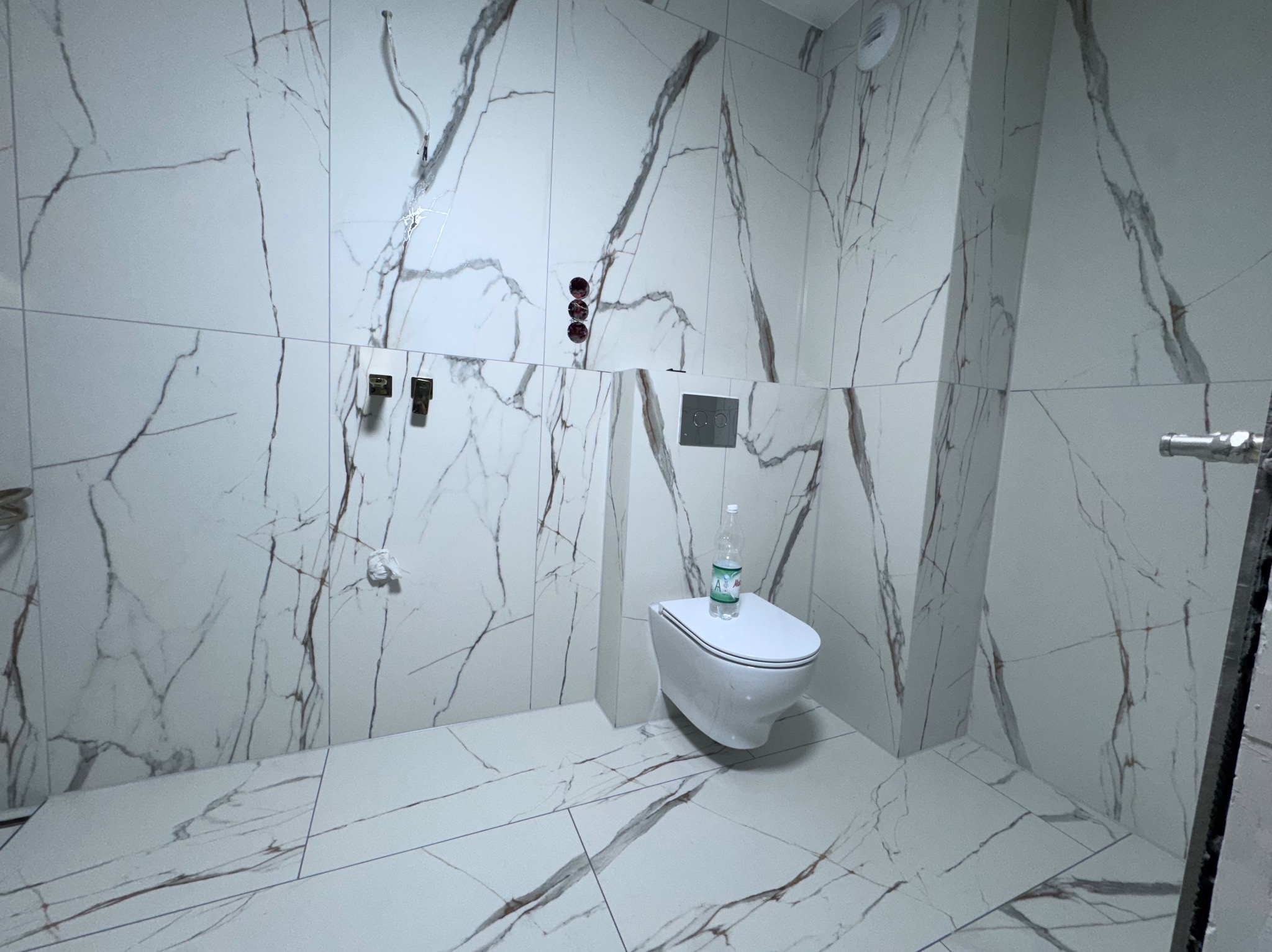 Biała łazienka z marmurowymi płytkami, wisząca toaleta i bateria podtynkowa. Widoczne przyłącza wodne i odpływowe. Minimalistyczny design z butelką wody na sedesie.