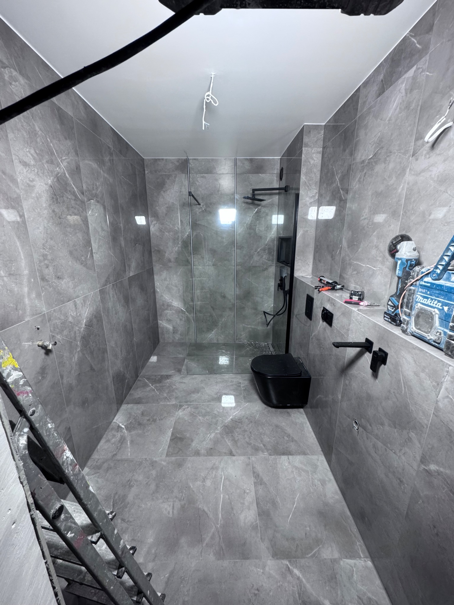 Szara łazienka po remoncie z kabiną prysznicową typu walk-in, czarną armaturą i wiszącą toaletą. Na ścianach i podłodze płytki imitujące marmur. Widoczne narzędzia.