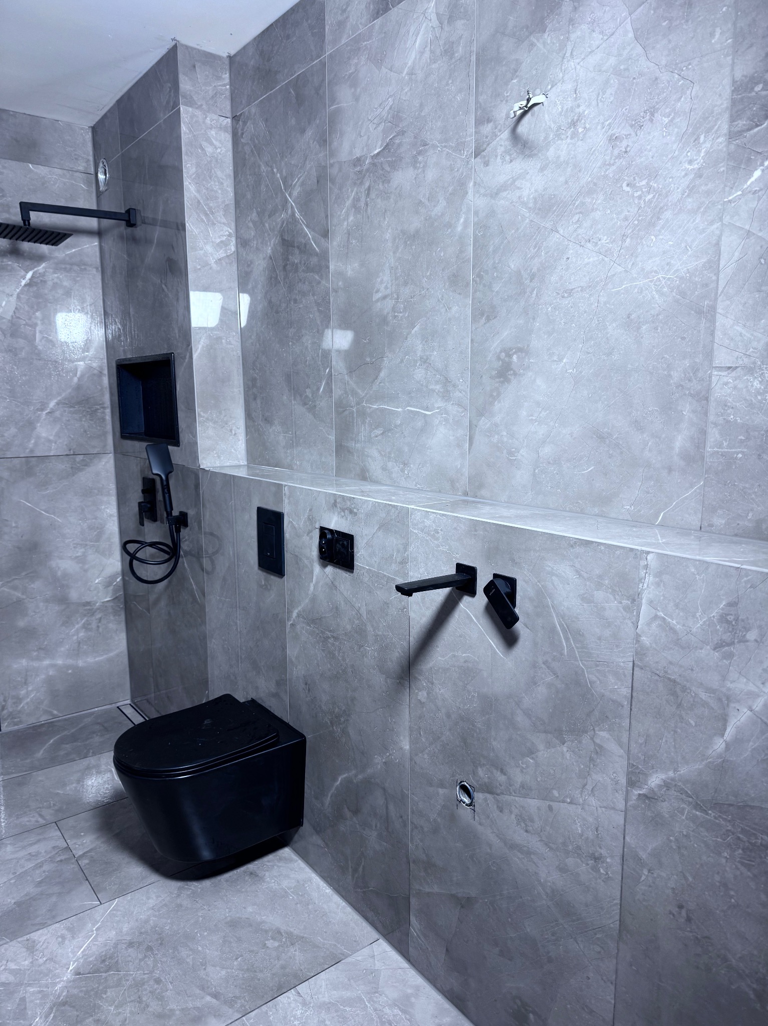 Nowoczesna łazienka w szarościach z czarną armaturą: wisząca toaleta, deszczownica, bateria umywalkowa. Minimalistyczny design, marmurowe płytki, oświetlenie punktowe.