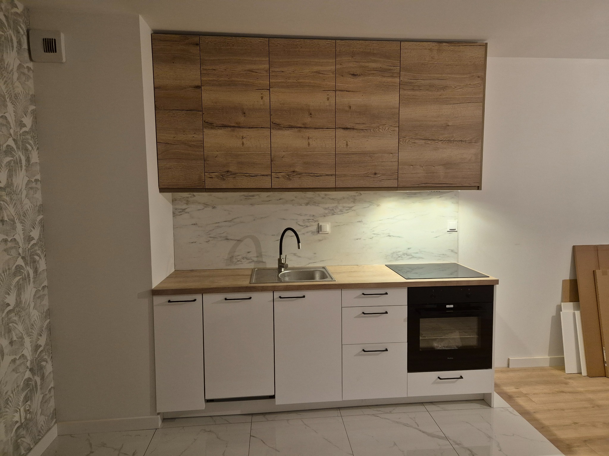 Nowoczesna kuchnia z białymi frontami szafek dolnych i drewnopodobnymi szafkami górnymi, marmurowy backsplash, czarna płyta indukcyjna i piekarnik, widoczne kartony po meblach.