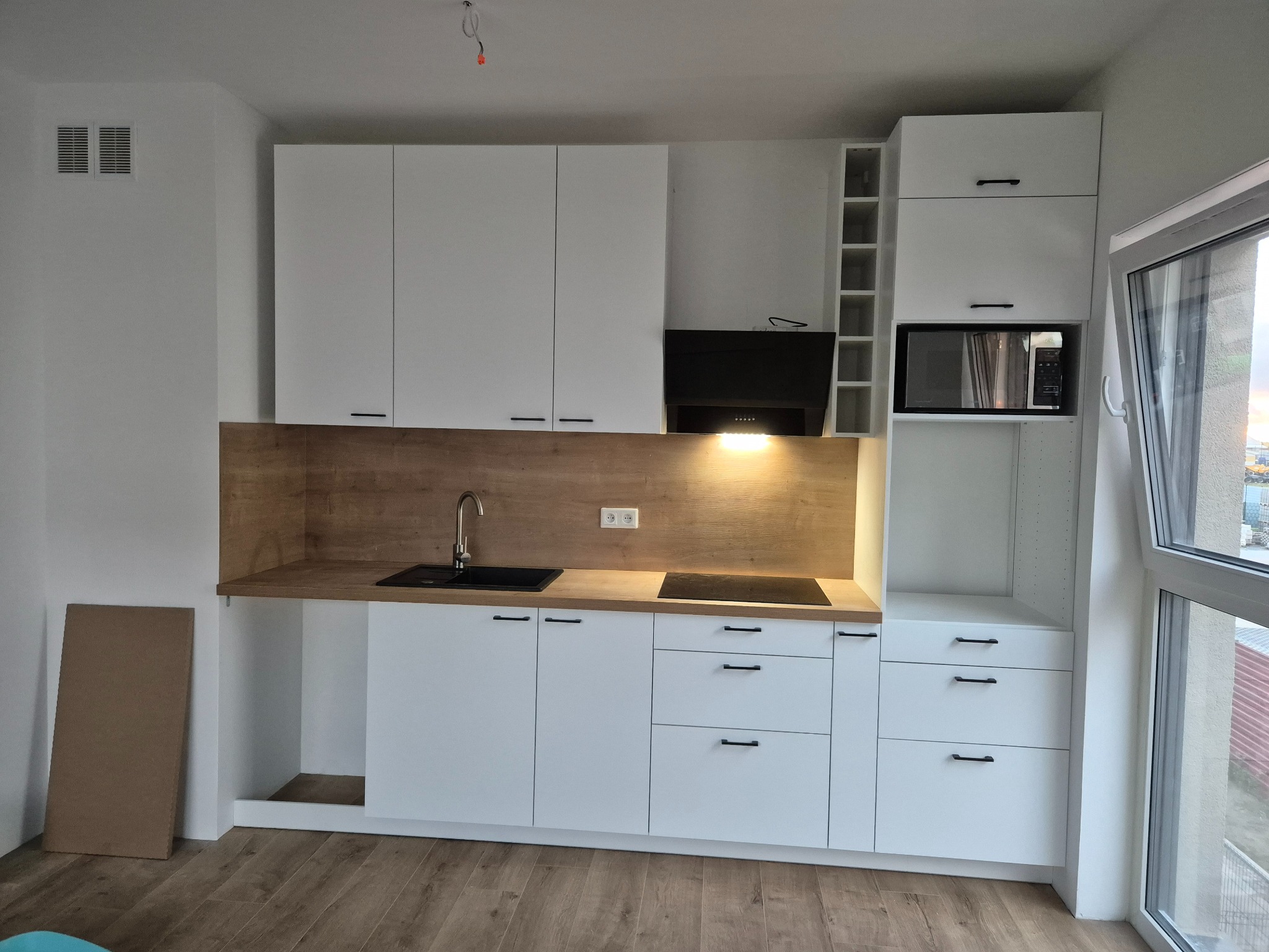 Nowoczesna kuchnia z białymi frontami szafek i drewnianym blatem, czarny zlew i płyta indukcyjna, minimalistyczne uchwyty, widok na okno i element niezamontowanej płyty bocznej.