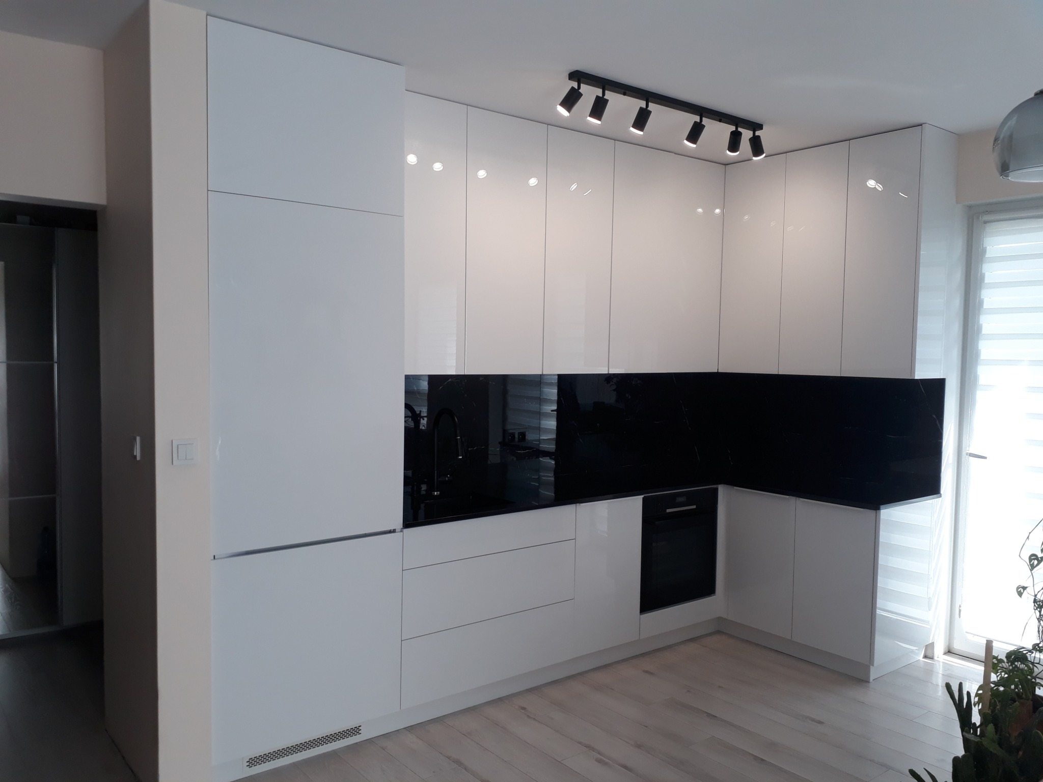 Nowoczesna, biała kuchnia na wymiar z czarnym blatem i panelem ściennym, oświetlona reflektorami sufitowymi.