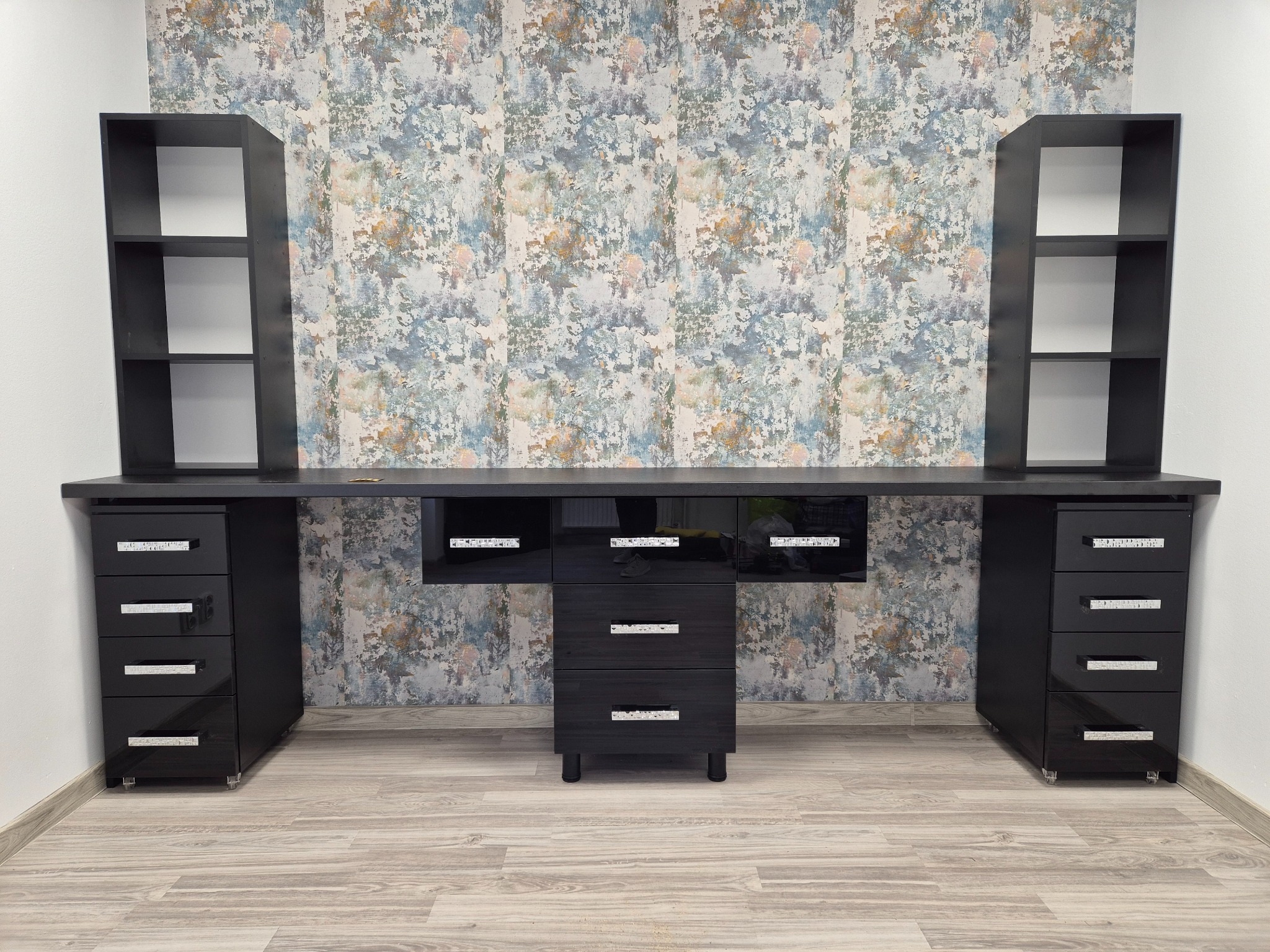 Czarny zestaw mebli biurowych z błyszczącymi uchwytami, składający się z biurka, szafek z szufladami na kółkach i otwartych półek, na tle wzorzystej tapety.