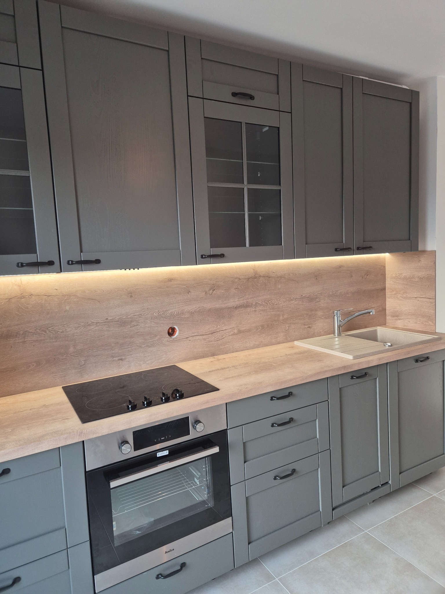Szara kuchnia z drewnianym blatem i oświetleniem LED podszafkowym, płyta indukcyjna i piekarnik Amica w zabudowie.