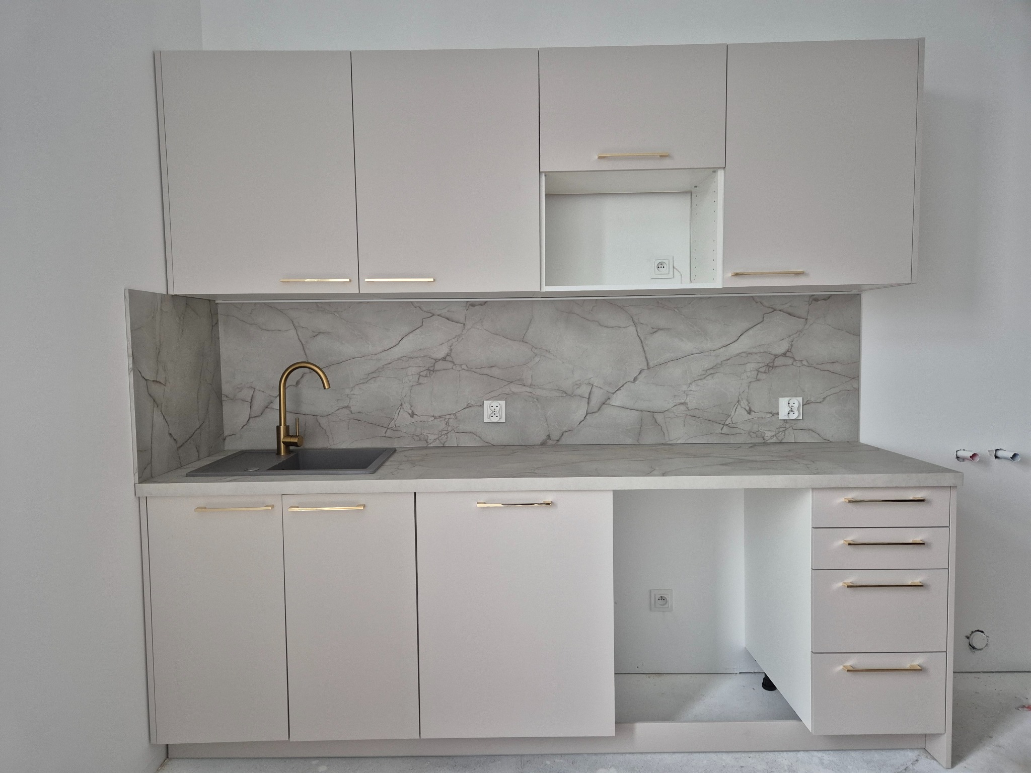 Jasna zabudowa kuchenna z marmurowym panelem, złotą armaturą i minimalistycznymi uchwytami, widoczne gniazdka elektryczne.