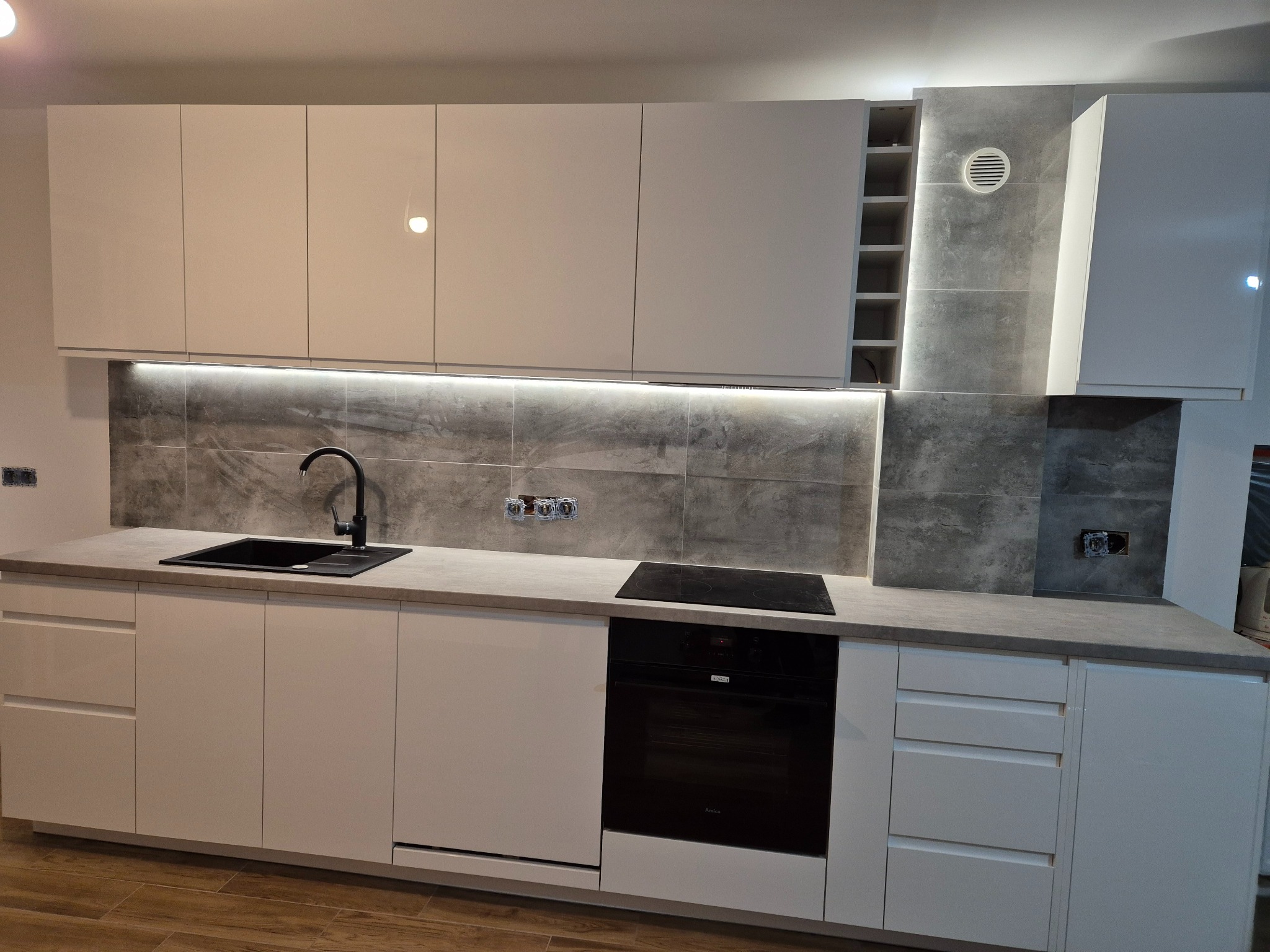 Nowoczesna kuchnia z białymi, lakierowanymi frontami, szarym blatem i płytkami imitującymi beton, czarny zlew i płyta indukcyjna, oświetlenie LED pod szafkami górnymi.