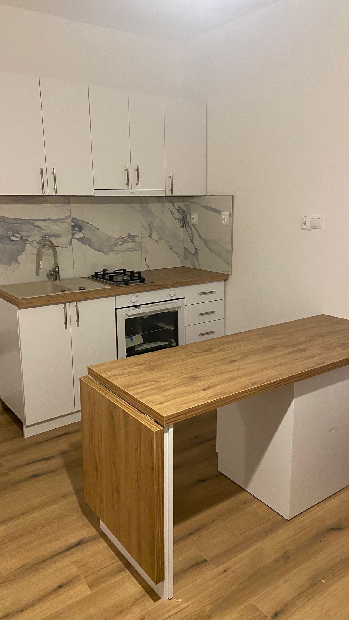 Nowoczesna kuchnia z białymi szafkami, marmurkowym backsplashiem i blatem z drewna w odcieniach brązu, z rozkładanym stołem/wyspą kuchenną.