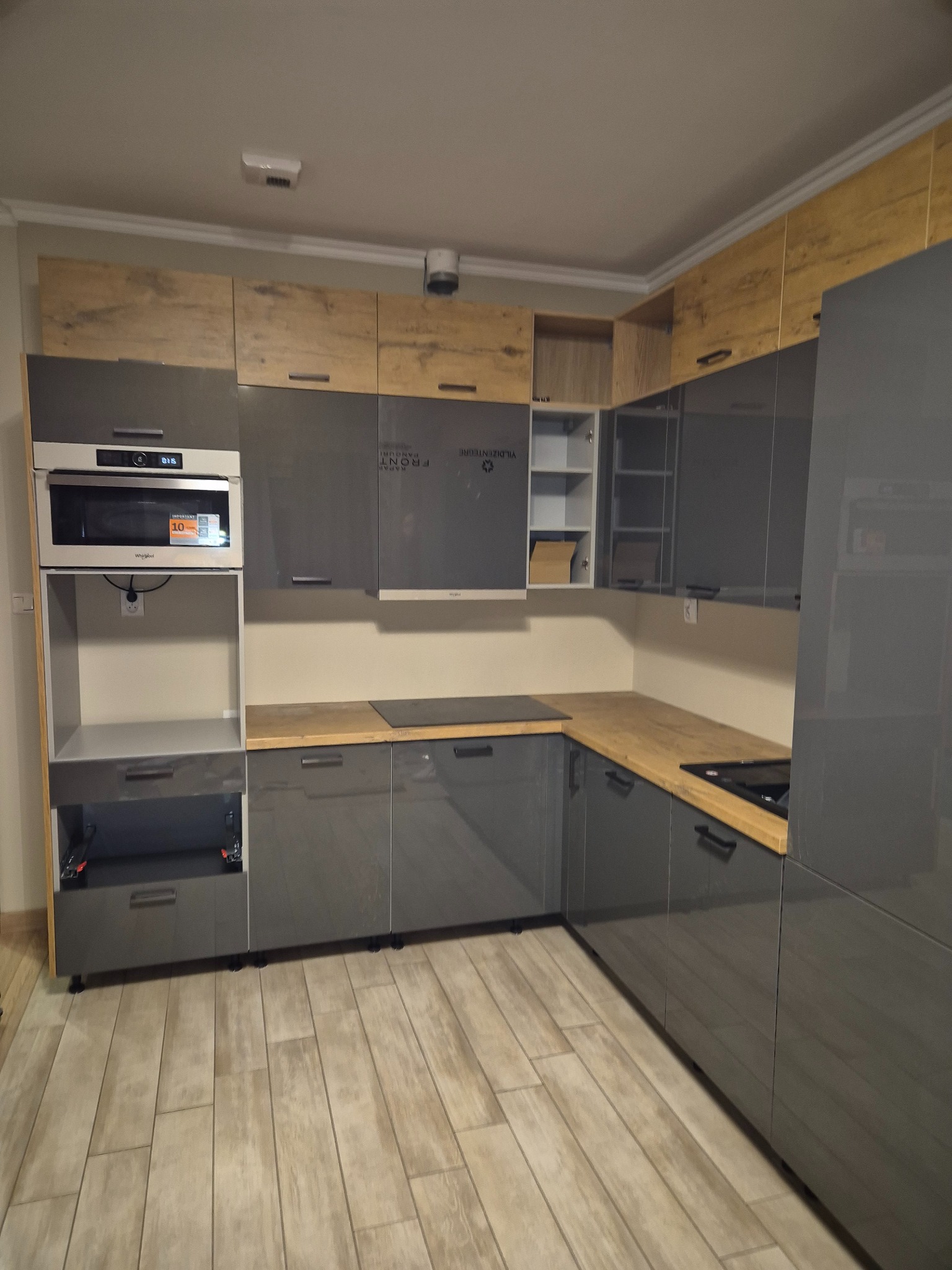 Szara kuchnia z drewnianymi akcentami, widoczne szafki górne i dolne, płyta indukcyjna w drewnianym blacie, otwarty dolny moduł z szufladami na prowadnicach.