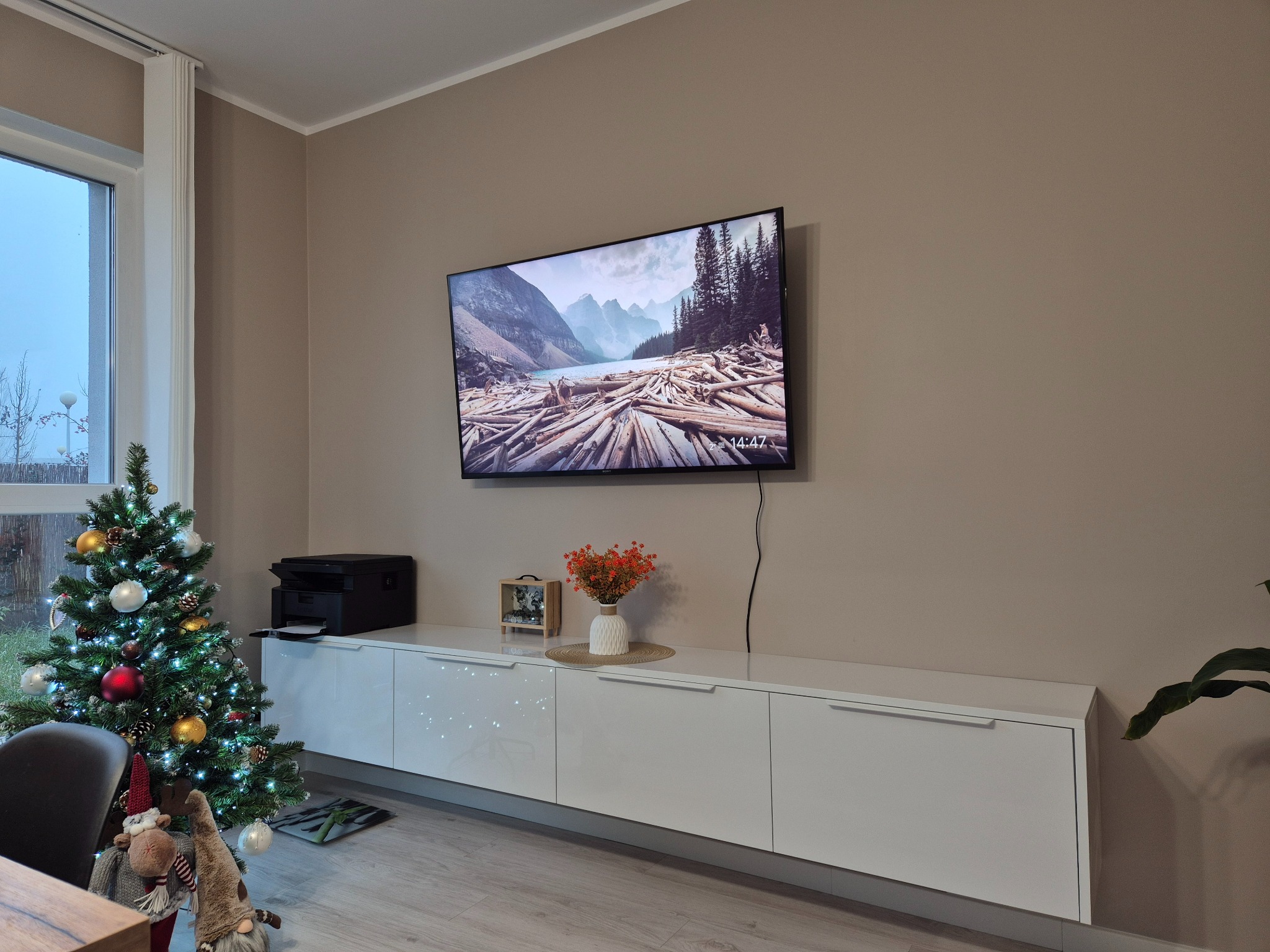 Biała szafka RTV z błyszczącymi frontami, na której stoi drukarka, ramka ze zdjęciem i wazon z pomarańczowymi kwiatami, a nad nią wisi telewizor; obok choinka z lampkami i ozdobami świątecznymi.