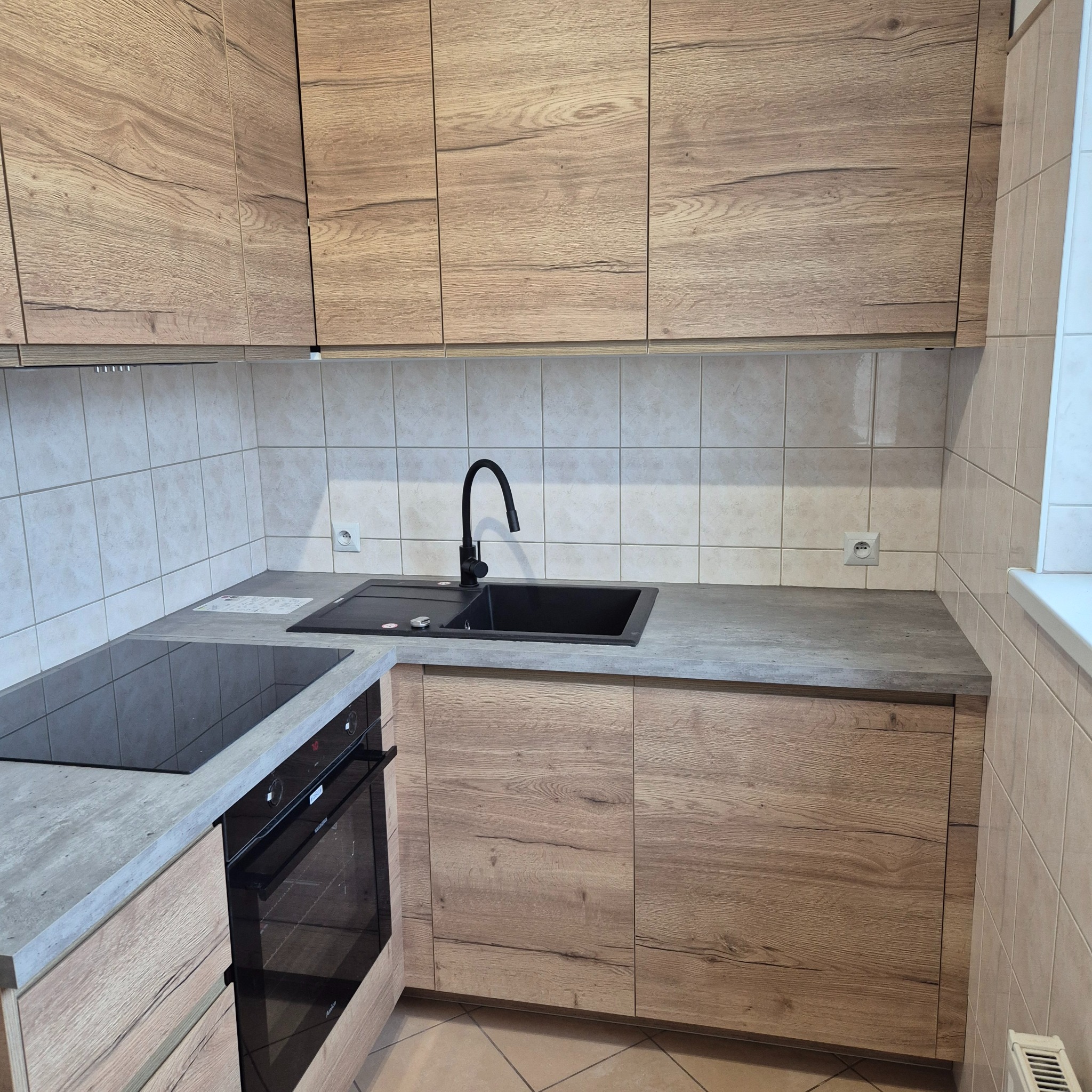 Nowoczesna kuchnia z drewnianymi frontami, czarnym zlewem i płytą indukcyjną, wykończona płytkami w odcieniach beżu.