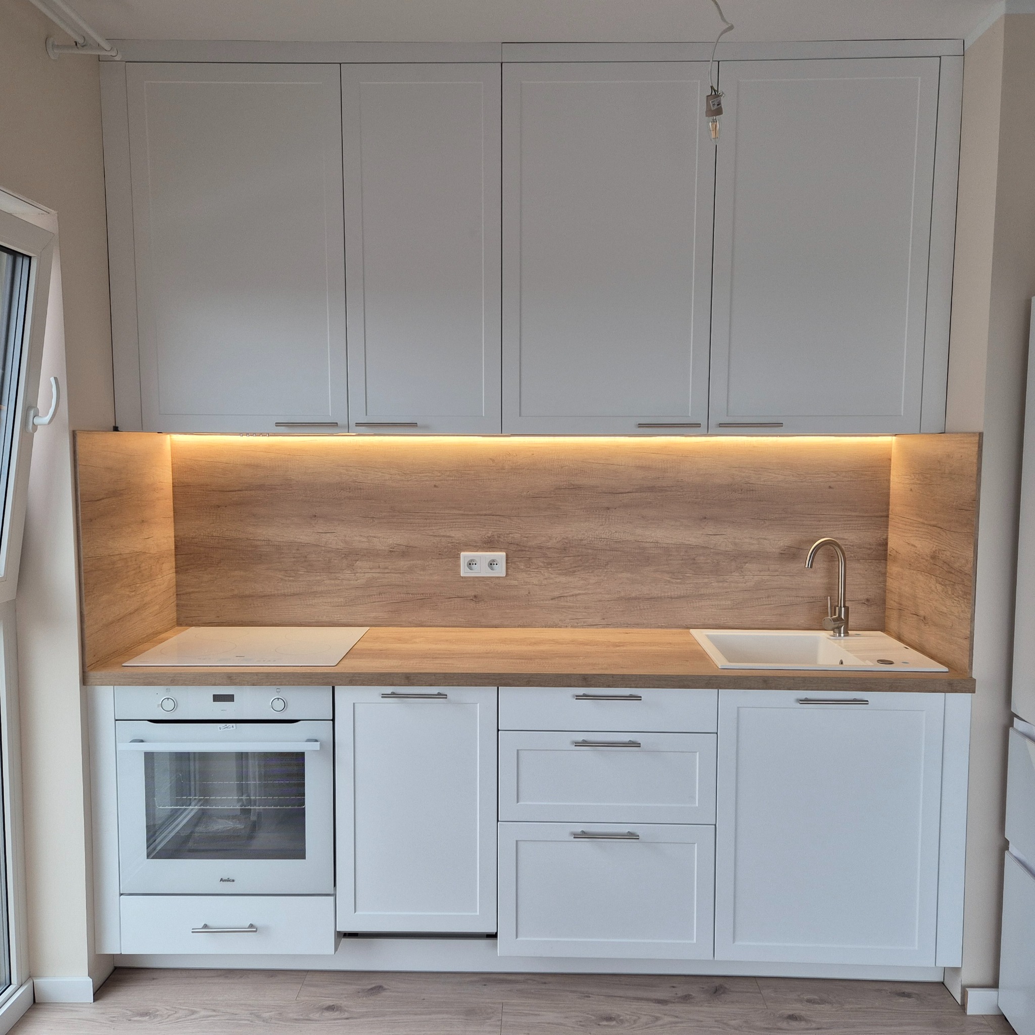 Nowoczesna, jasna kuchnia z białymi szafkami i drewnianym blatem, oświetlona listwą LED pod szafkami wiszącymi, z widoczną białą płytą grzewczą i piekarnikiem Amica.