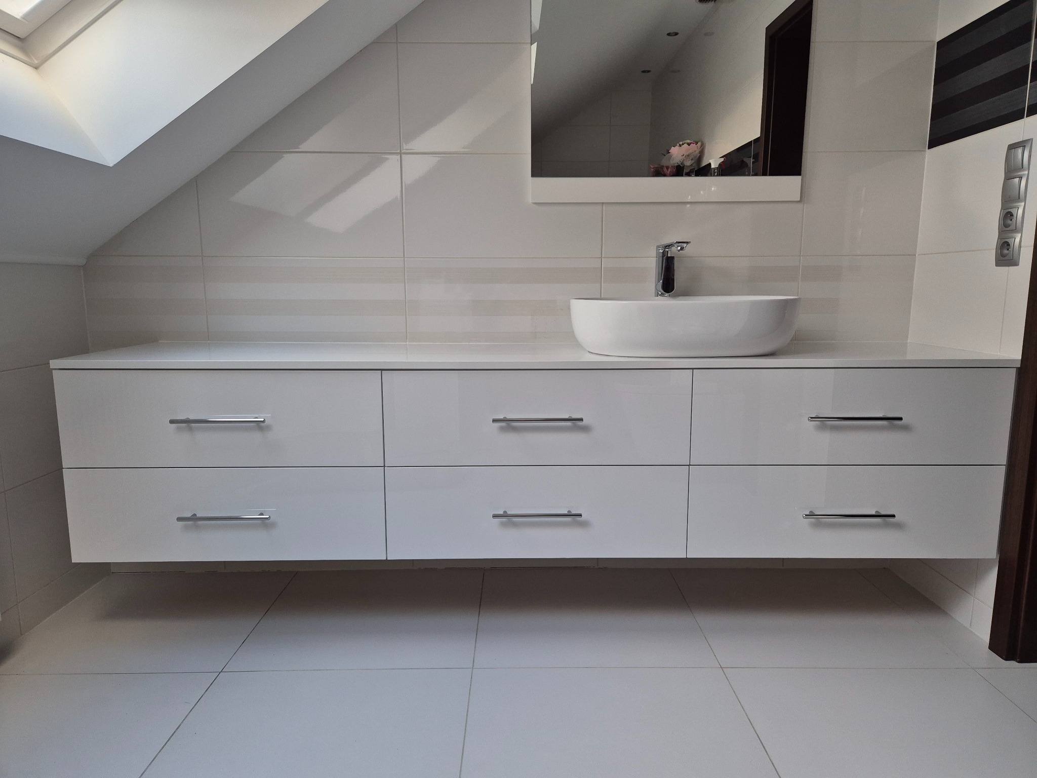 Biała, wisząca szafka łazienkowa z sześcioma szufladami i umywalką nablatową w Pobiedziskach, zamontowana pod skosem dachu.