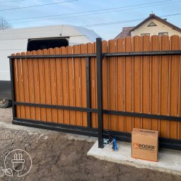 DAVCAM DAWID LEŻOŃ - Brama przesuwna z brązowymi panelami i czarną ramą, automat Roger Technology obok. Biały bus w tle. Nowoczesny design bramy wjazdowej.