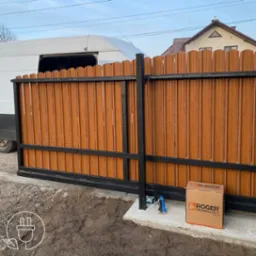 Brama przesuwna z brązowymi panelami i czarną ramą, automat Roger Technology obok. Biały bus w tle. Nowoczesny design bramy wjazdowej.