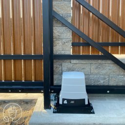 DAVCAM DAWID LEŻOŃ - Zamontowany automat do bramy przesuwnej na betonowej podstawie, widoczna listwa zębata i fragment brązowej bramy z czarną ramą na tle kamiennego muru.