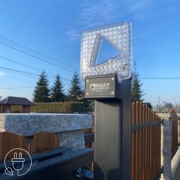 DAVCAM DAWID LEŻOŃ - Lampa LED Roger Technology na słupku bramy wjazdowej, z widocznym logo producenta i elementami automatyki bramy. Błękitne niebo w tle, drzewa iglaste.