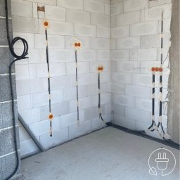 DAVCAM DAWID LEŻOŃ - Wnętrze w trakcie prac elektrycznych: w ścianach z betonu komórkowego poprowadzone kable, widoczne puszki instalacyjne. Czarny kabel biegnie wzdłuż ściany.