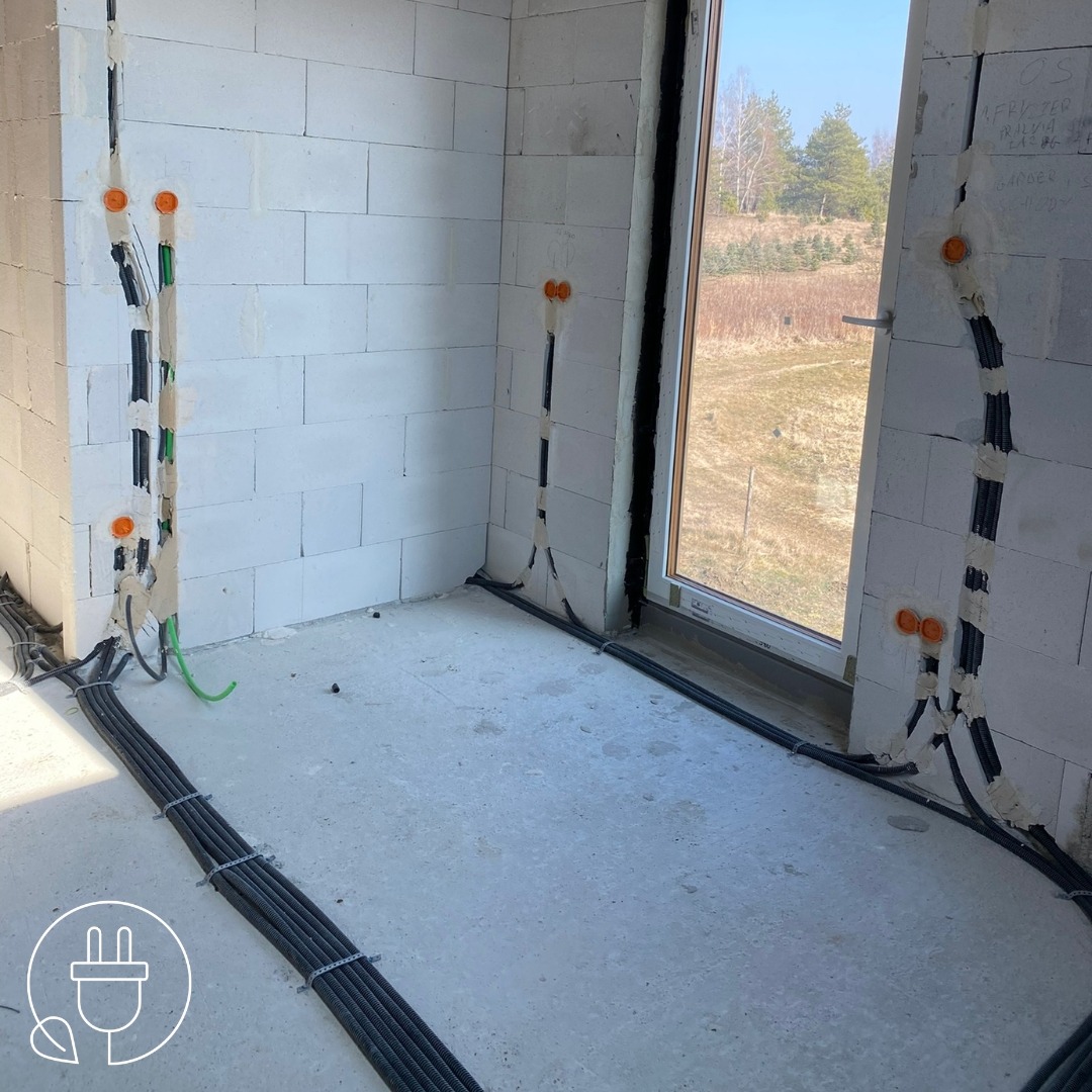 Wnętrze w trakcie prac elektrycznych: kable w peszlach prowadzone w bruzdach ścian z betonu komórkowego, puszki instalacyjne, okno z widokiem na zewnątrz.