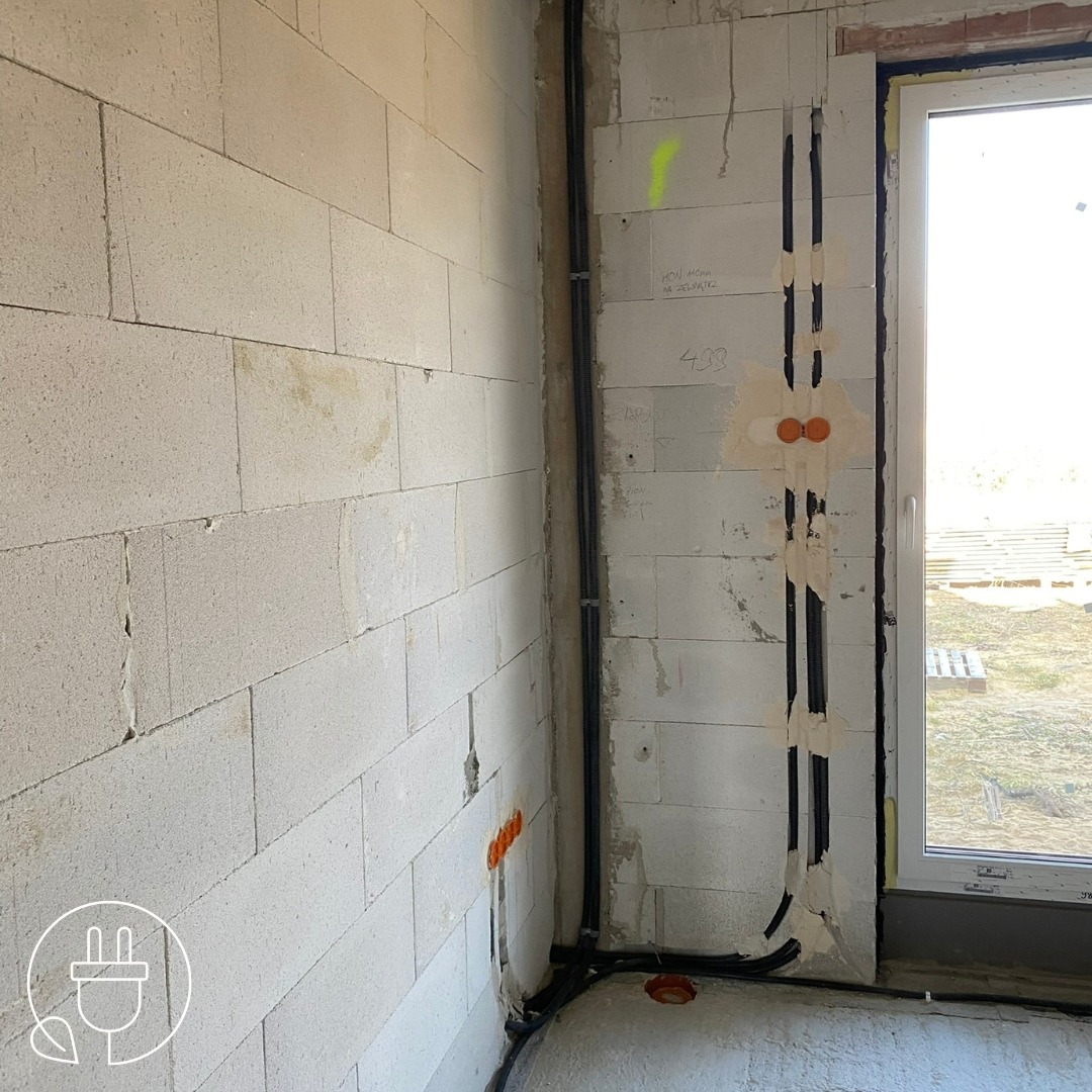 Wnętrze w trakcie prac elektrycznych: widoczne pionowe kable w ścianie z betonu komórkowego, zabezpieczone taśmą, oraz otwory pod gniazdka. Okno w tle.