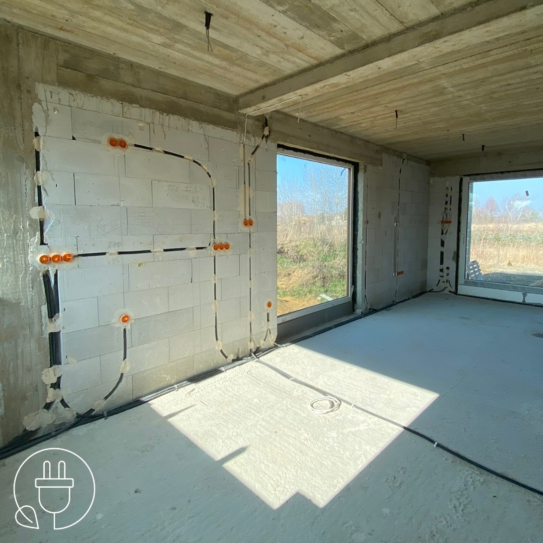 Surowy stan: instalacja elektryczna wewnątrz budynku z białego betonu komórkowego, widoczne okna i kable w peszlach. Gniazdka elektryczne w kolorze pomarańczowym.