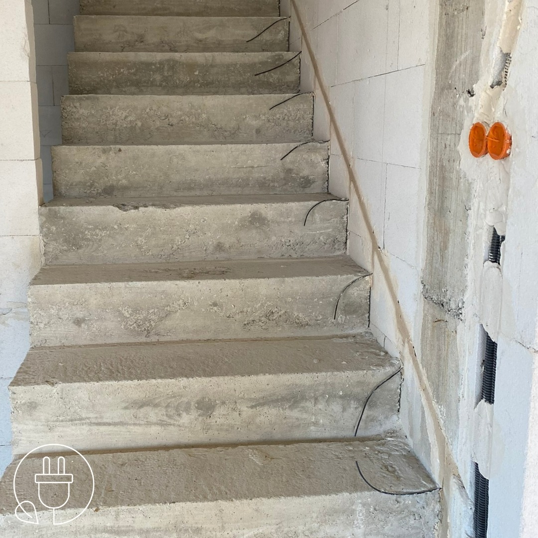 Surowe betonowe schody w trakcie budowy z wystającymi kablami elektrycznymi i puszkami pod gniazdka w ścianie z betonu komórkowego.