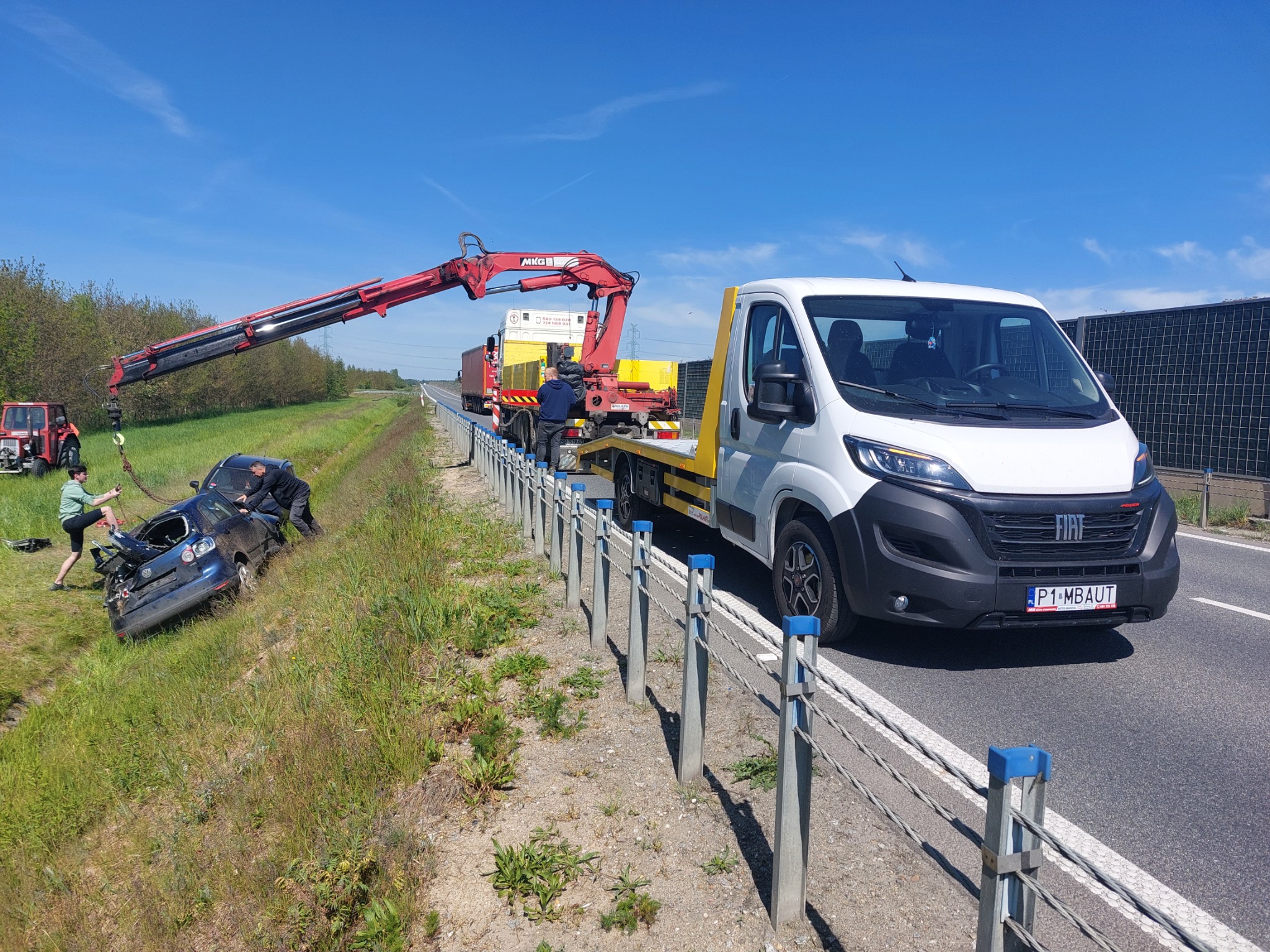 Uszkodzony, granatowy samochód osobowy wyciągany z rowu za pomocą dźwigu zamontowanego na lawecie, obok stoi biały Fiat Ducato z żółtą platformą lawety, na pierwszym planie barierka ochronna.
