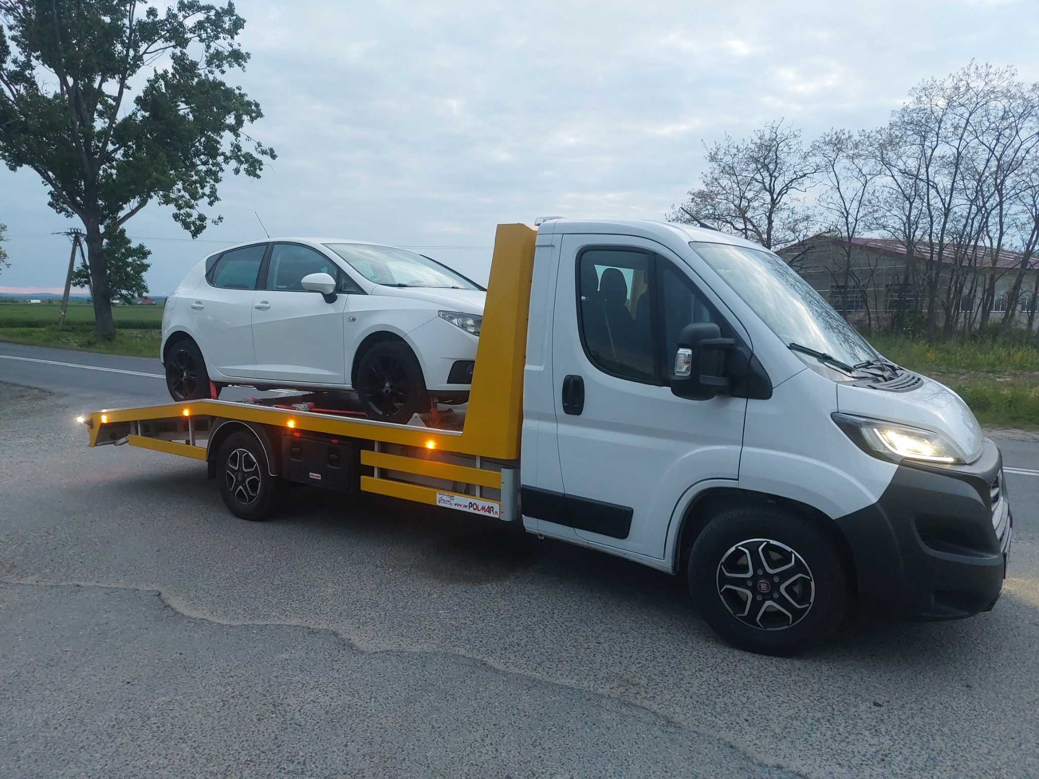 Biała laweta z załadowanym białym samochodem osobowym marki Seat Ibiza z czarnymi felgami, zaparkowana na poboczu drogi w pochmurny dzień.