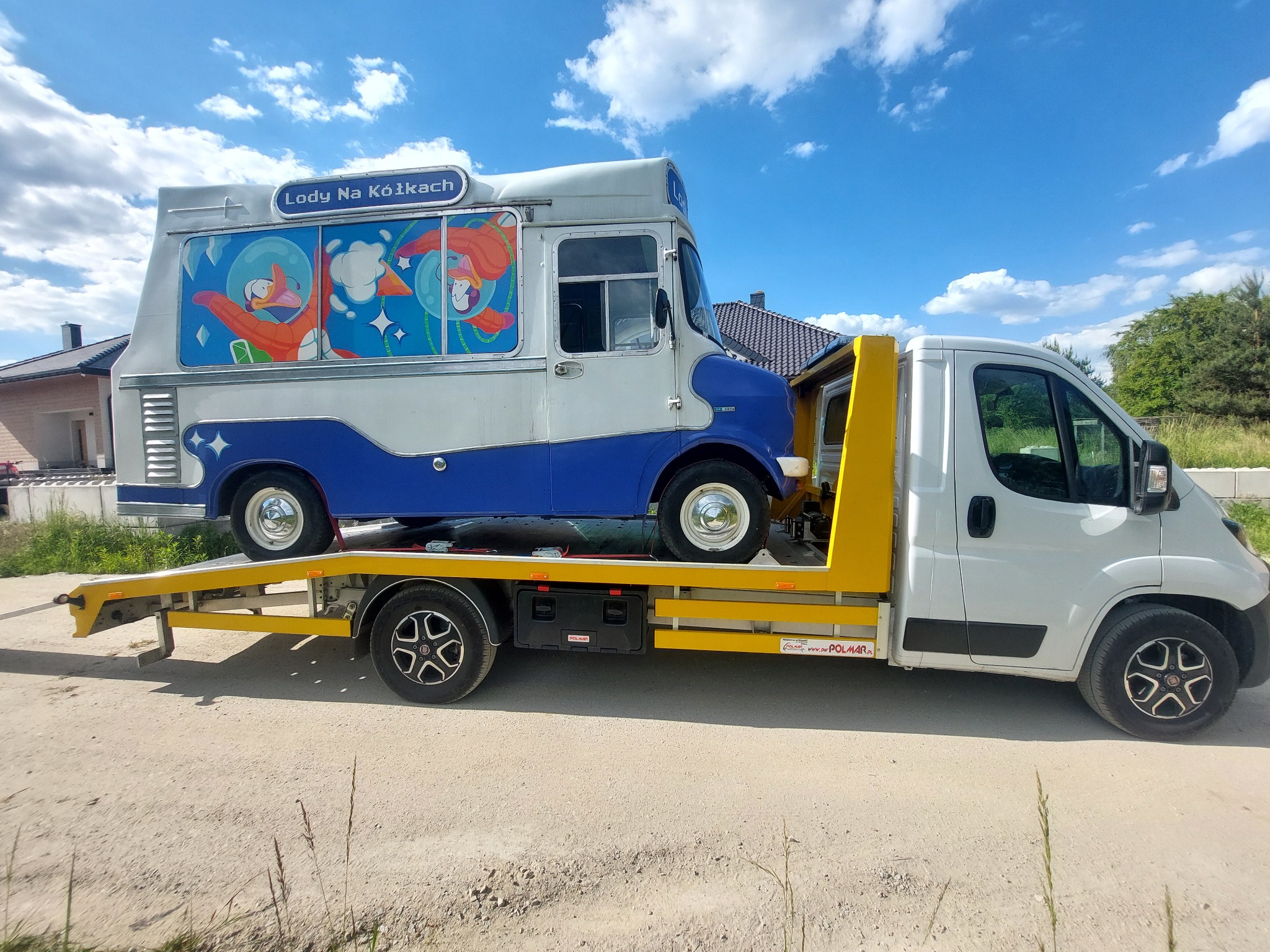Laweta przewozi food trucka z napisem 'Lody Na Kółkach' i rysunkami kaczek w kosmicznych skafandrach.