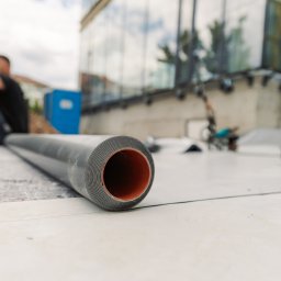 Qubik Dachy Jakub Pulkowski - Rozwinięta rolka czarnej membrany dachowej na białym podłożu, z widocznym budynkiem w tle. Detal na strukturze materiału i pomarańczowym wnętrzu rolki.