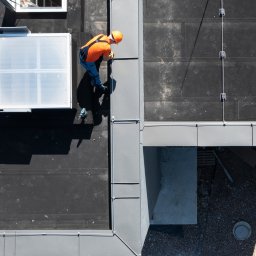 Qubik Dachy Jakub Pulkowski - Praca na dachu z papy termozgrzewalnej. Dekarz w pomarańczowej koszulce i kasku montuje obróbkę blacharską przy świetliku dachowym. Widok z góry, narzędzia obok.