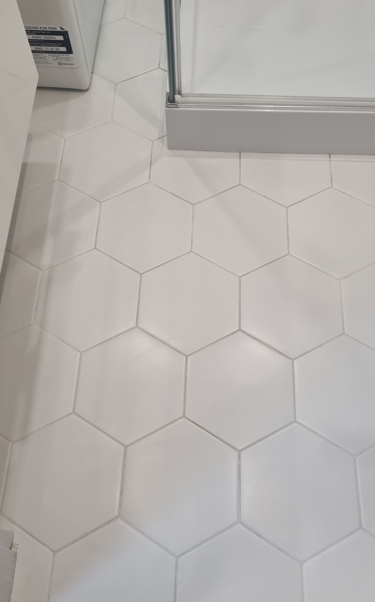 Białe, heksagonalne płytki podłogowe w łazience, widoczne fragmenty kabiny prysznicowej i pralki. Minimalistyczny design, czyste fugi, jasne oświetlenie.