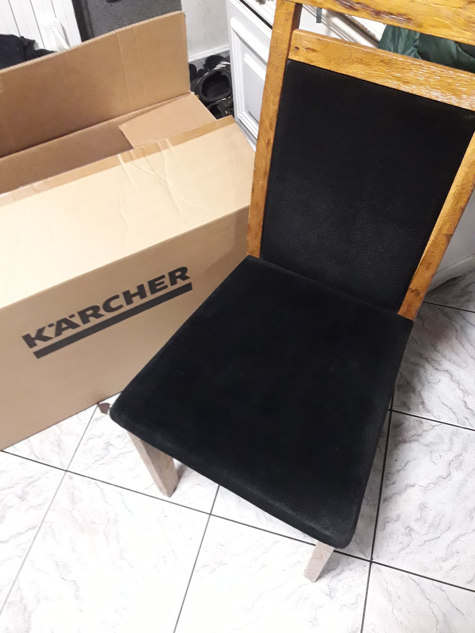 Krzesło z czarną tapicerką i drewnianą ramą stoi obok kartonu Karcher na marmurowej podłodze. Widoczne detale tapicerki i struktura drewna.